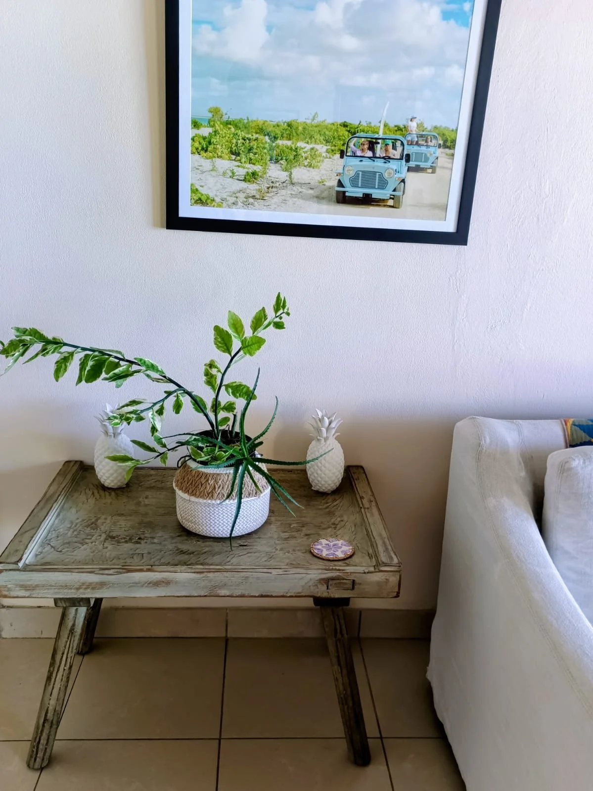 Interior of a room with a wooden table holding a potted plant with long green leaves, two ceramic pineapple decorations, and a small decorative item. A framed photograph of people riding in and driving golf carts on a sandy path hangs on the wall above the table. Part of a white armchair is visible on the right.