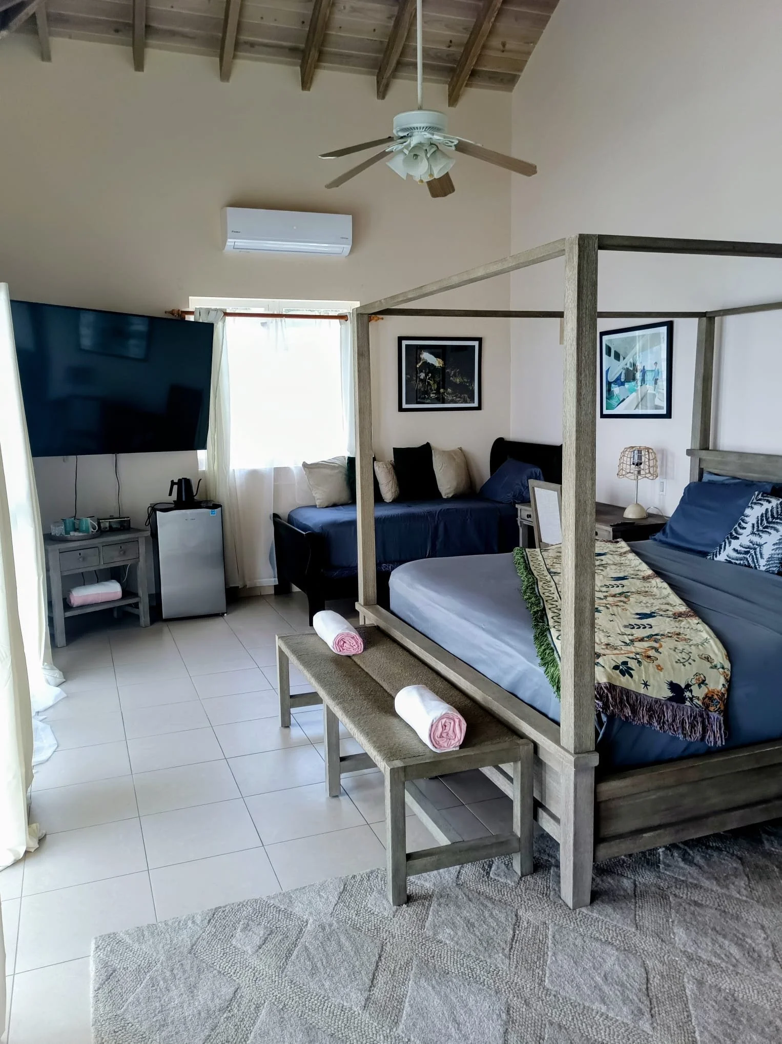 A cozy bedroom with a four-poster bed with blue bedding and a colorful throw blanket, two pink rolled towels on a small bench, a gray nightstand with a lamp and decorations, a wall-mounted TV, framed pictures, a window with white curtains, and a ceiling fan with wooden blades.