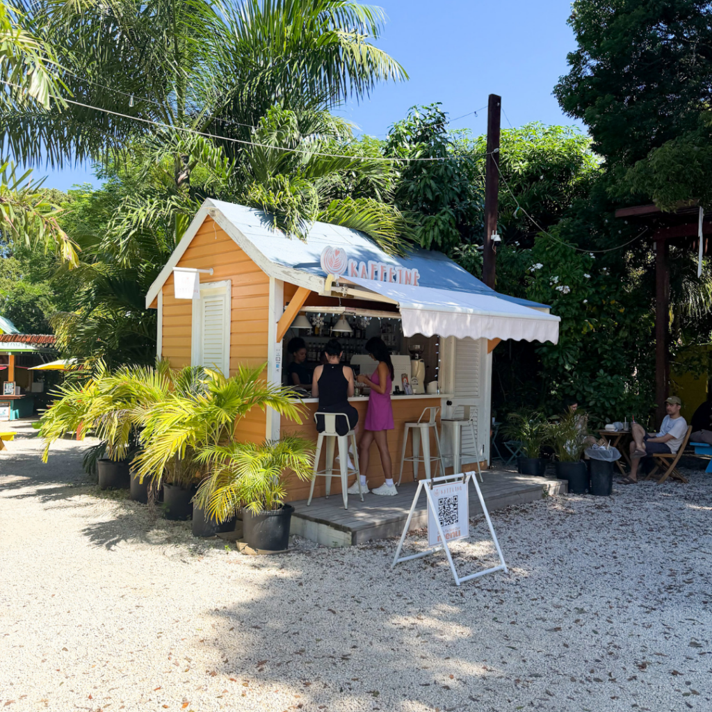 Small yellow cafe stand with white trim, surrounded by green tropical plants, with a chalkboard sign in front, and a couple of women ordering drinks inside, shaded by a trees with bright blue sky in the background.