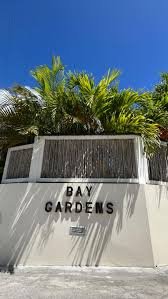 Entrance sign for Bay Gardens with tropical plants and a clear blue sky in the background.