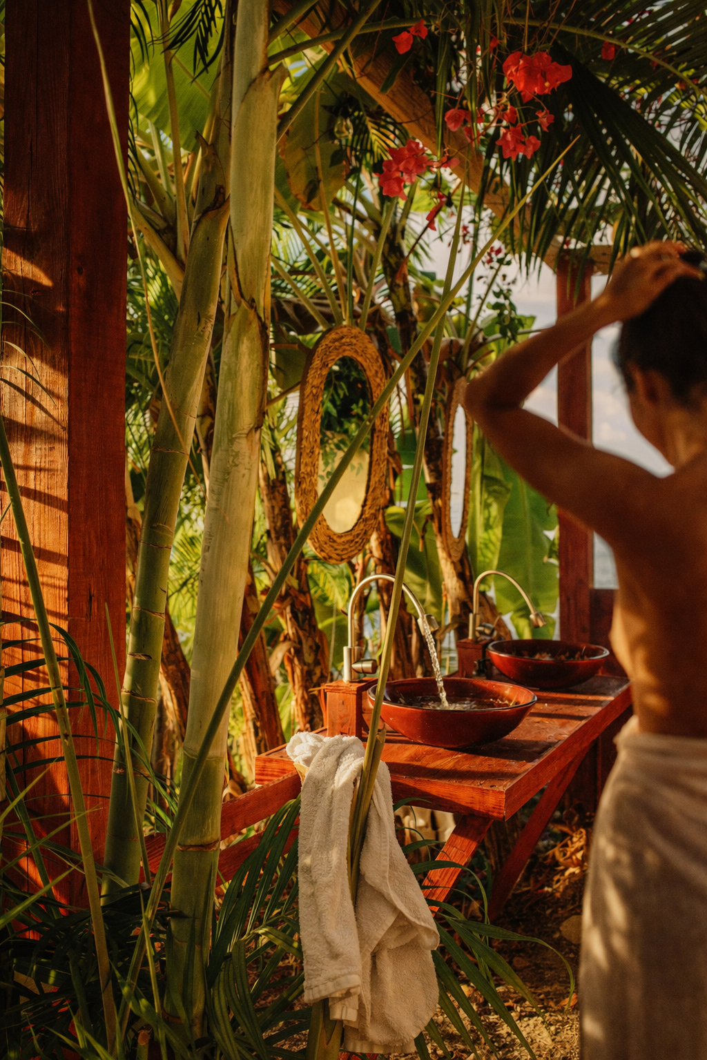 A person standing at an outdoor bathroom sink with two brown bowls, surrounded by lush green plants and hanging red flowers.