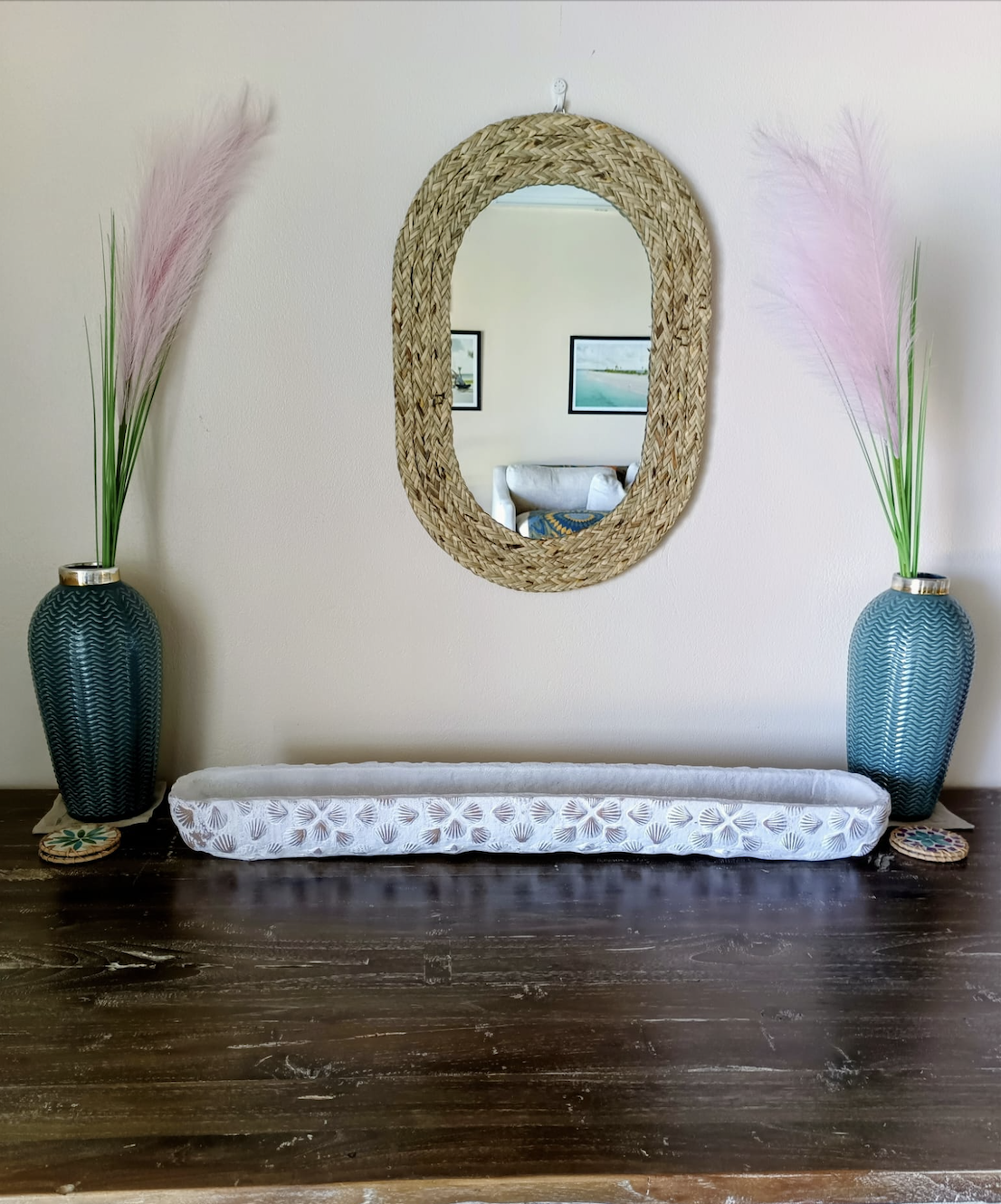 Decorative wall mirror with a woven frame, flanked by two tall blue vases with pink pampas grass on a dark wooden surface, in a minimalist indoor setting.