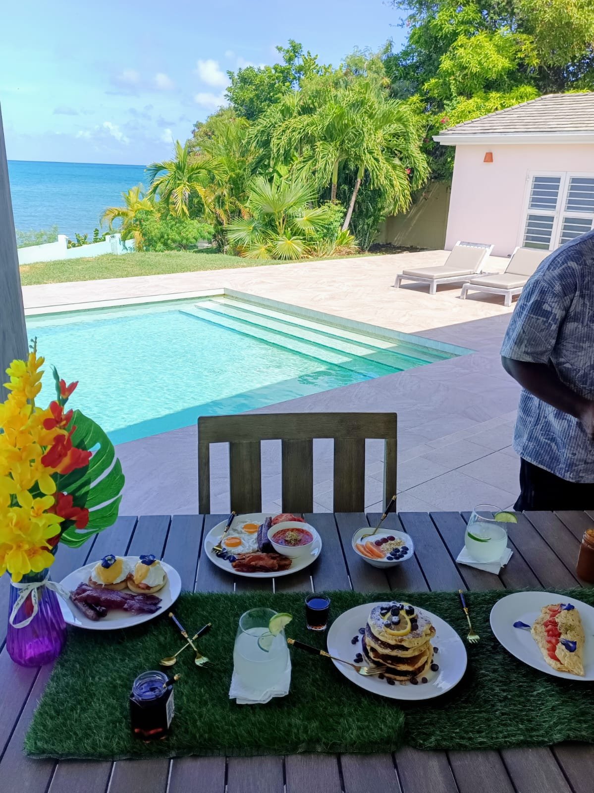 A poolside setup with a table of breakfast foods including pancakes, eggs, fruits, and drinks, overlooking a tropical landscape with palm trees and the ocean in the background.