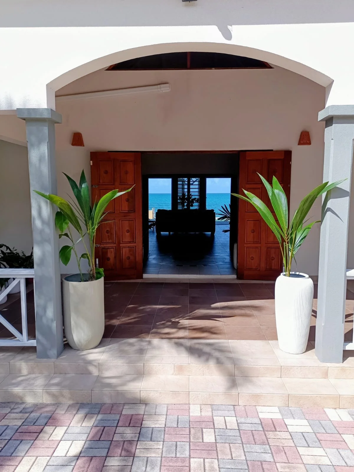 View of a patio leading to a living room with wooden double doors open to a beach scene with blue ocean and sky beyond. Potted plants on either side of the entrance, tiled flooring, and a white arch framing the doorway.