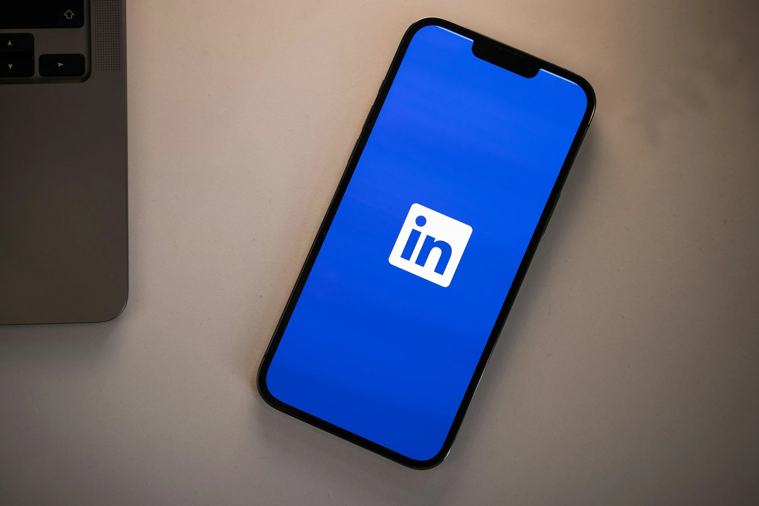 Un smartphone affichant le logo de LinkedIn sur son écran, posé sur une surface beige, à côté d'un ordinateur portable
