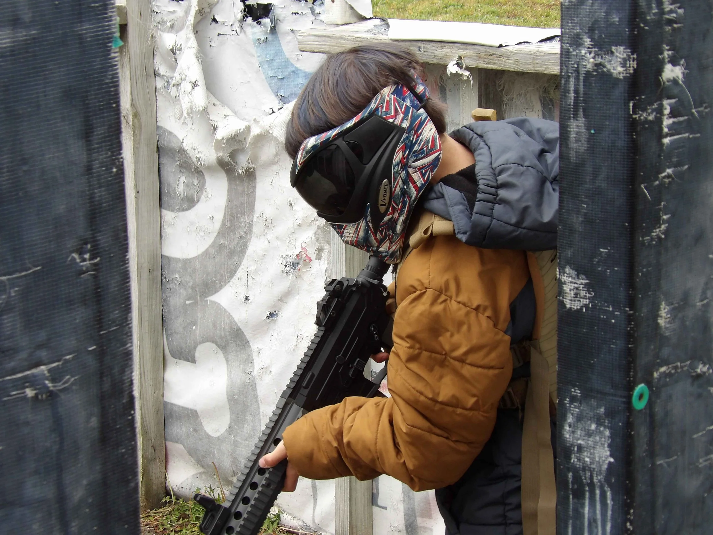 A person wearing a black and red helmet and a brown jacket holding a black rifle, positioned behind a wooden fence with black netting on the sides, in front of a weathered wall with peeling paint.