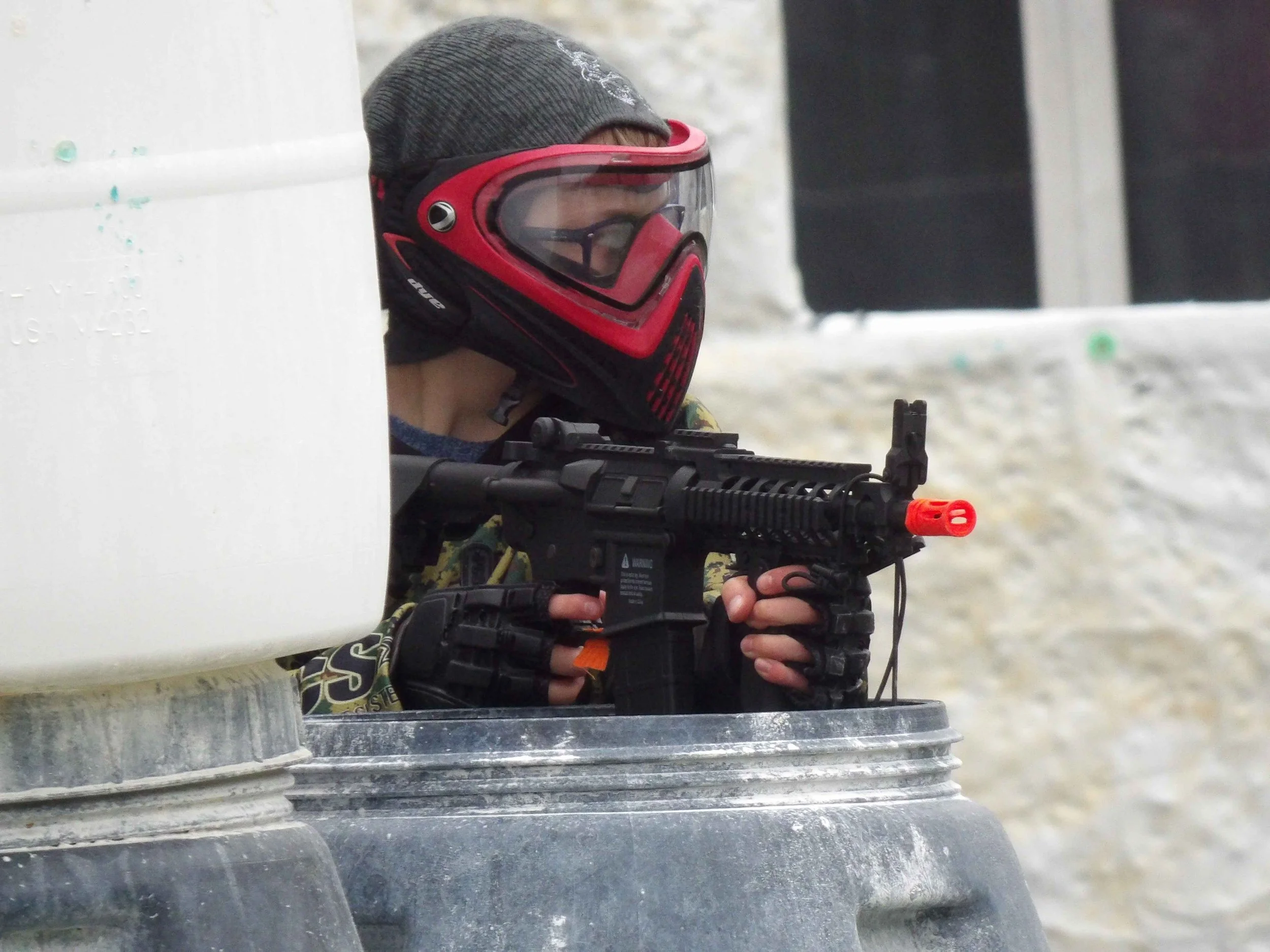 A person wearing a gray beanie, red and black protective goggles, and camouflage clothing is peering around a white object while holding a gun, possibly participating in a shooting activity.