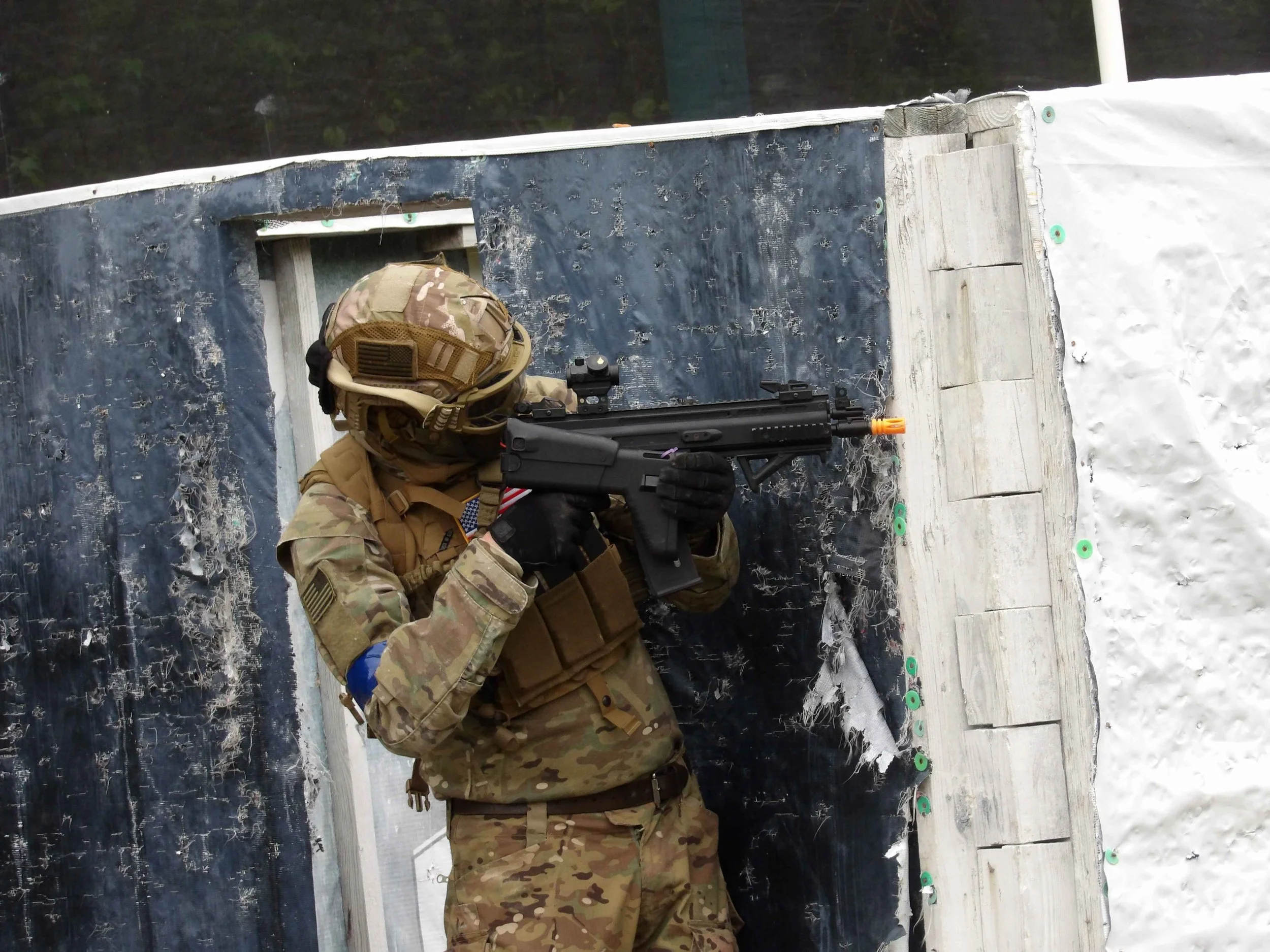 A soldier in camouflage uniform aiming a black and orange toy gun around a corner of a wooden and black structure in an outdoor winter setting with snow on the ground.