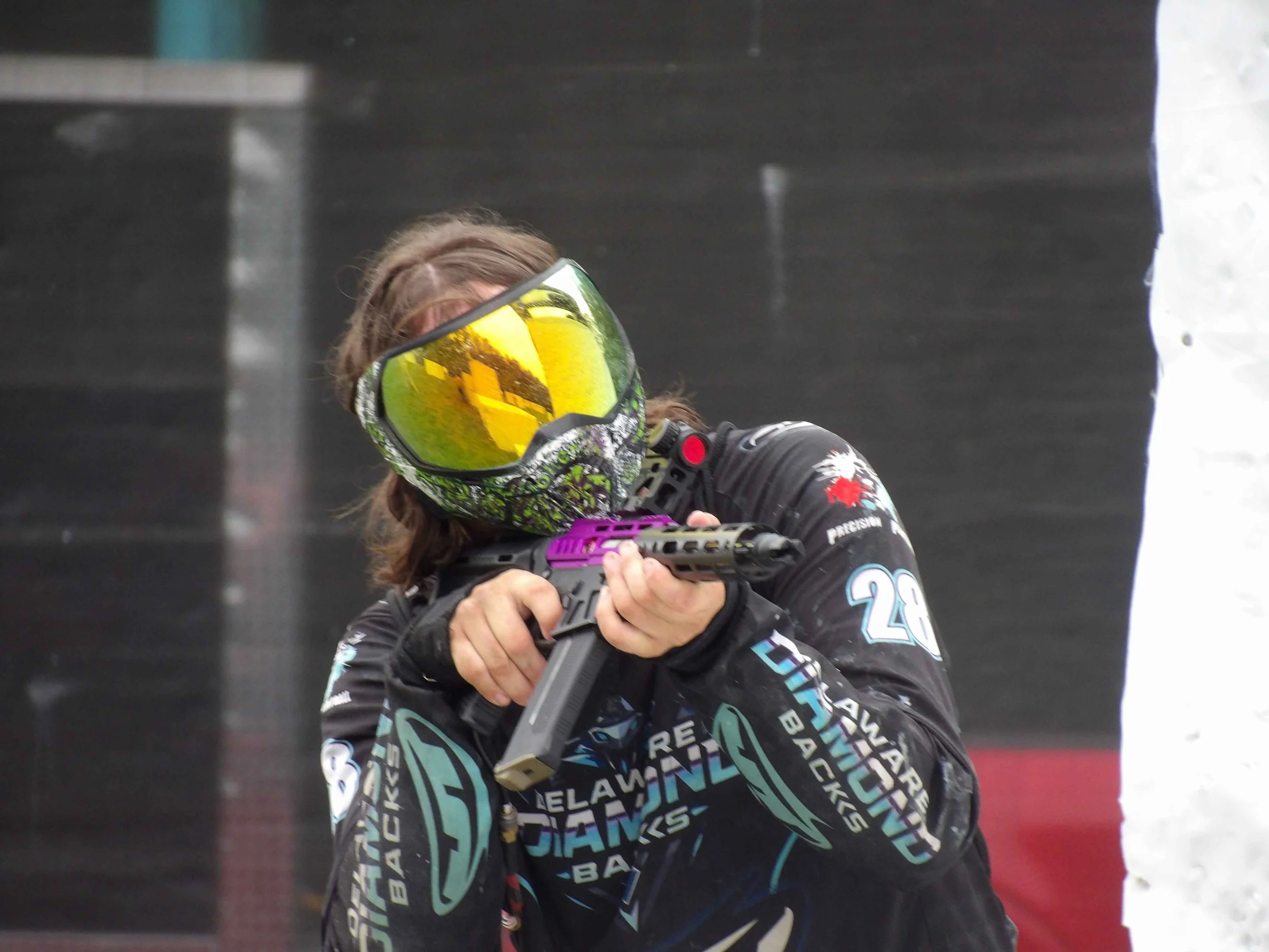 A person wearing a neon yellow and black ski or motocross helmet with a reflective yellow visor, black gloves, and a black jacket with colorful text and patterns aiming a gun through a shooting range or similar indoor setting.