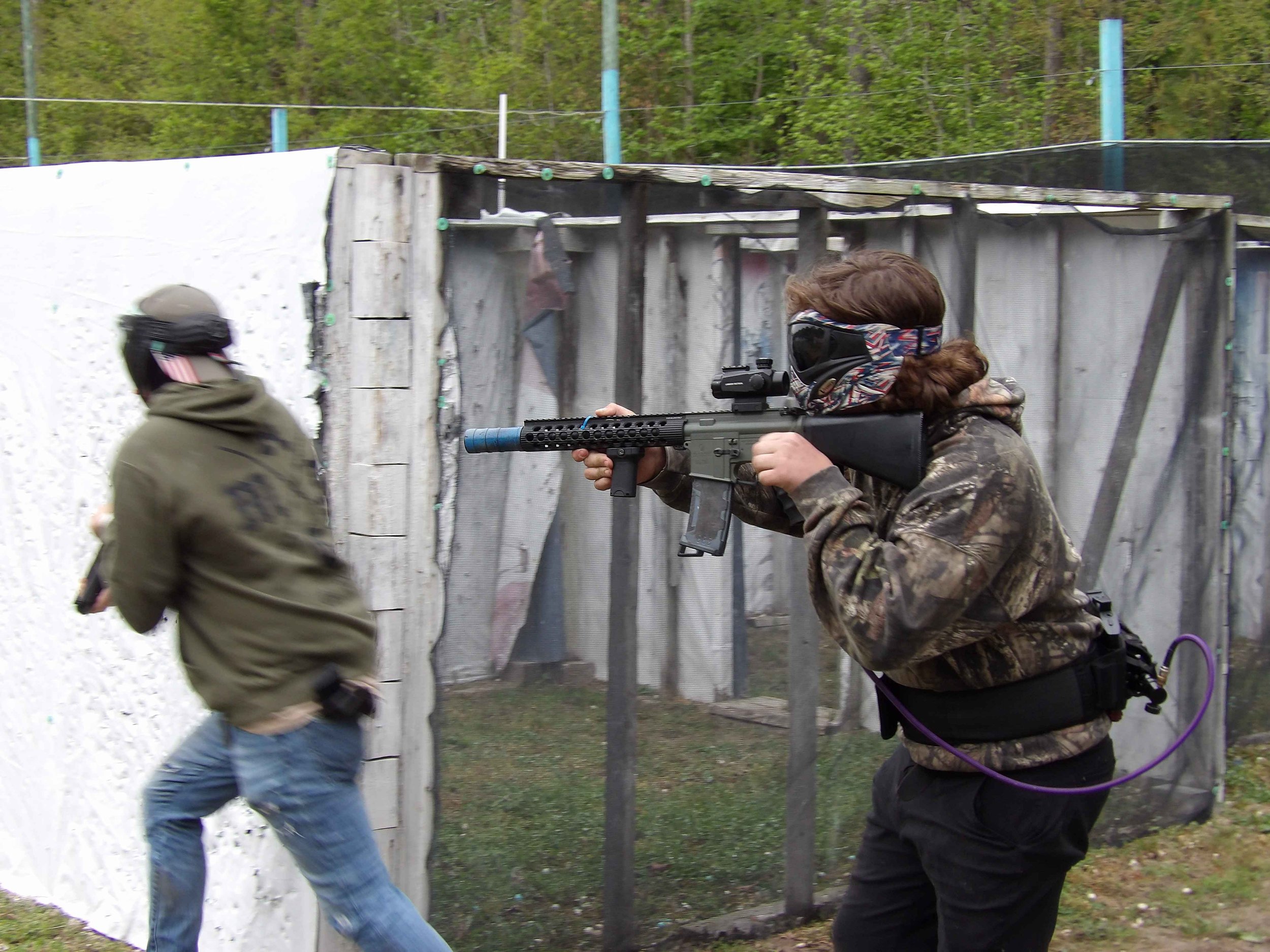A person in camouflage jacket and black pants holding a firearm with a suppressor, aiming at a shooting range wall. Another person in a green hoodie and blue jeans is walking past in the background.
