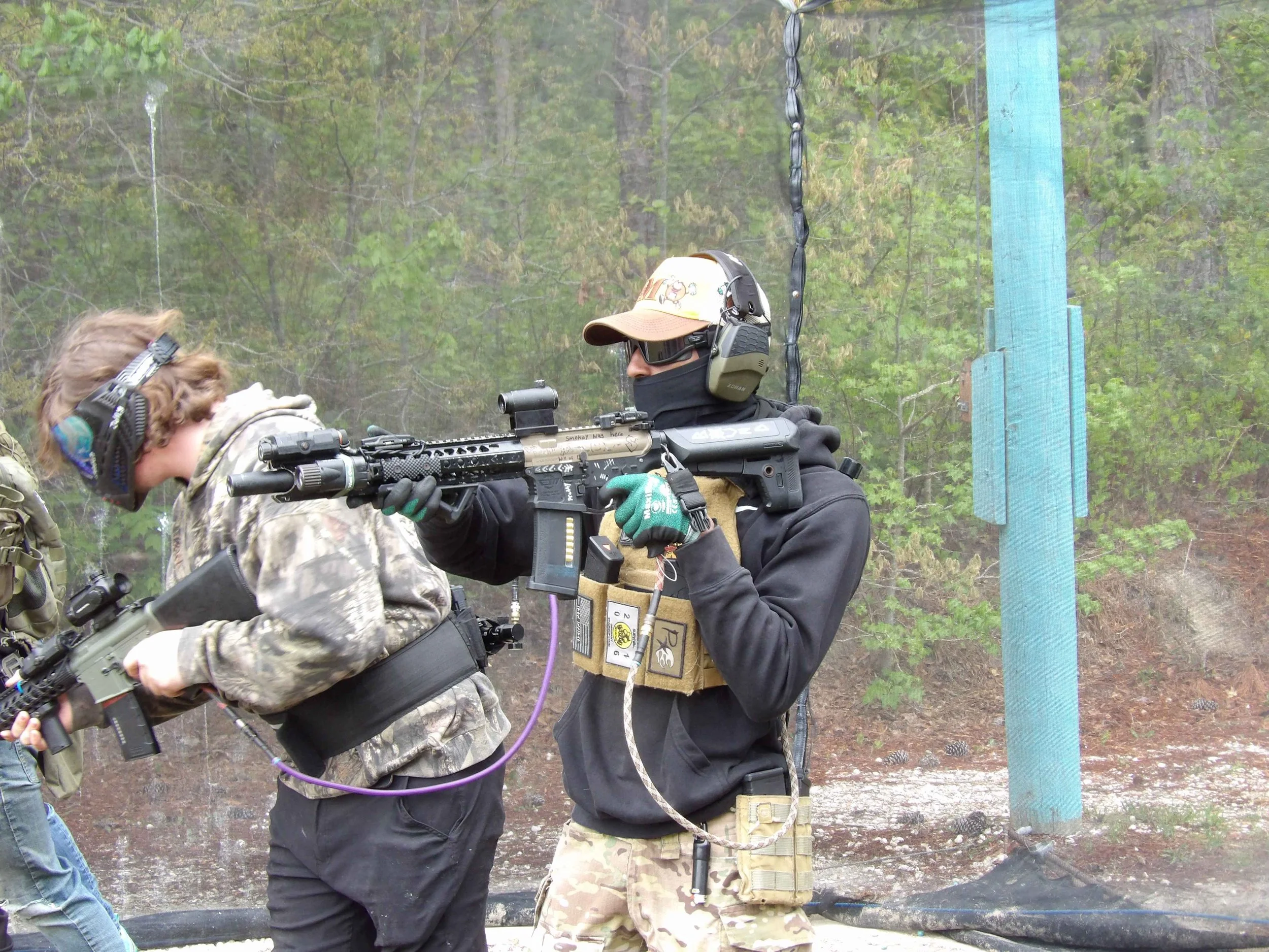 Two people in military gear aiming rifles at a shooting range in an outdoor setting with trees in the background. One person is wearing a cap, goggles, and headphones, while the other is in camouflage clothing with headphones.