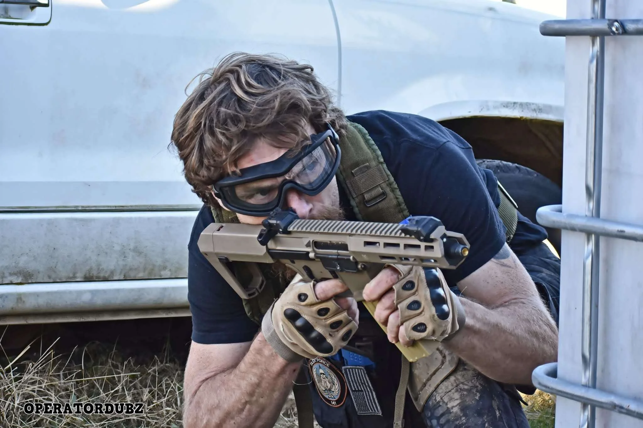 A man wearing tactical gear, protective goggles, and gloves crouches behind a vehicle, aiming a tan-colored handgun.