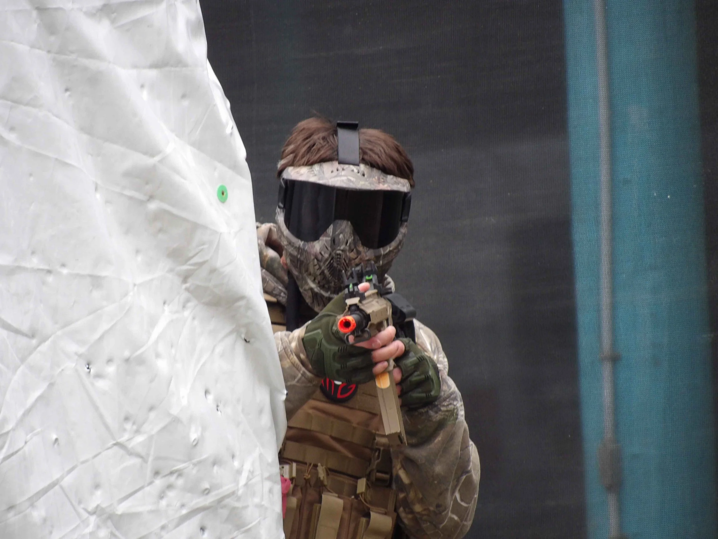 A person in camouflage military gear aiming a toy gun behind a large white rock with a security marker, in what appears to be an indoor laser tag or military simulation game.