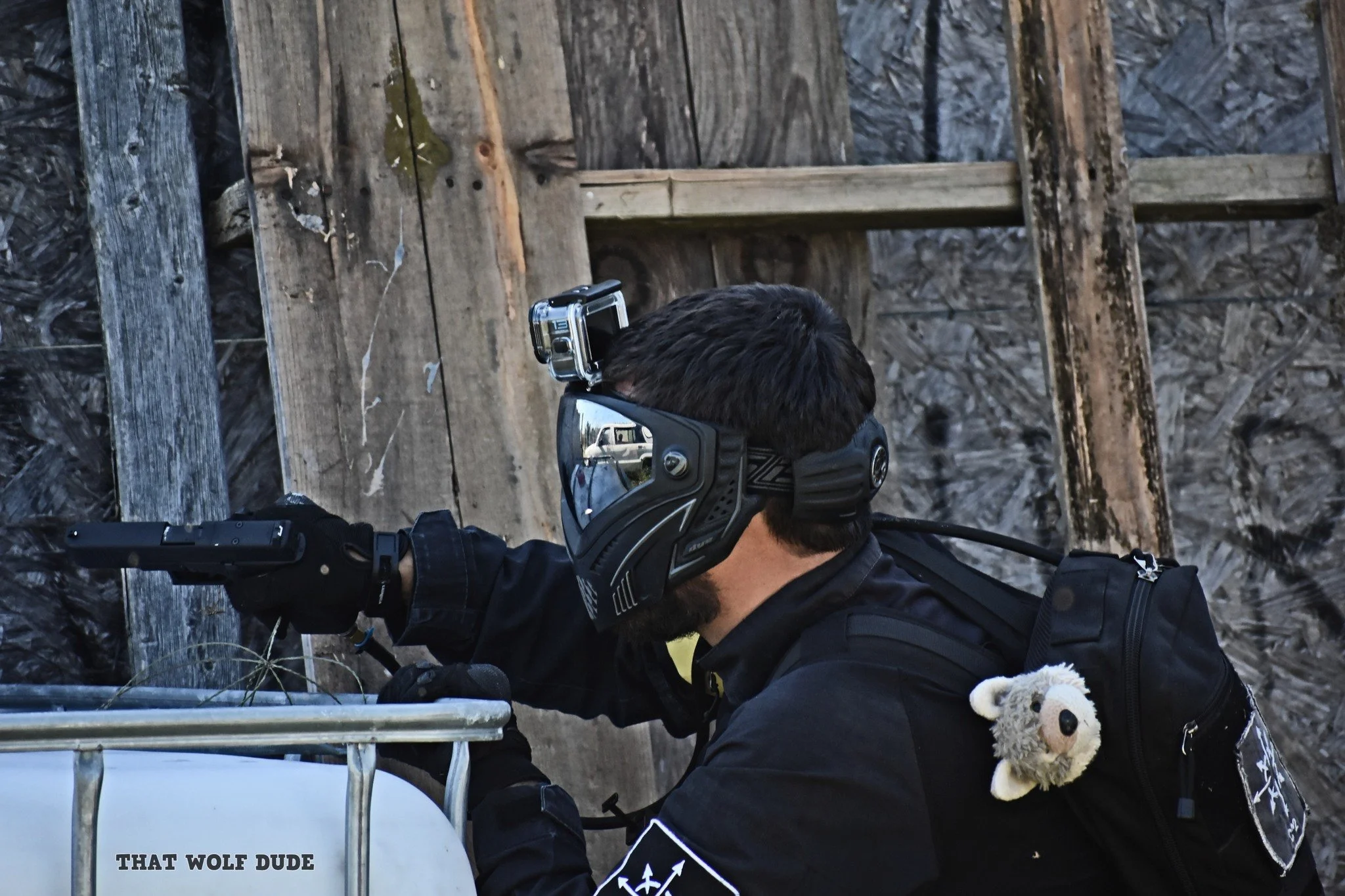 A person wearing a helmet with goggles, a black backpack with a small plush toy, black gloves, and black clothing, appears to be updating a metal cart or container labeled 'That Wolf Dude' in front of a wooden fence or structure.