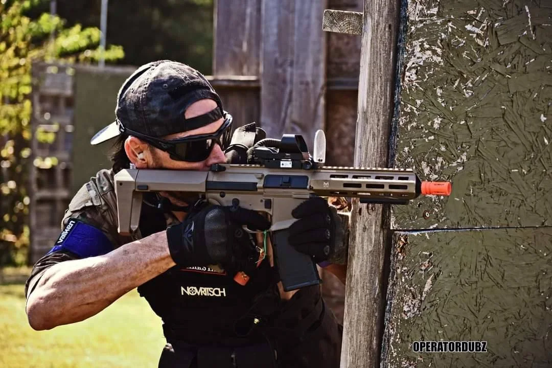 A person dressed in tactical gear, wearing sunglasses and a cap backward, aiming a rifle with an orange tip around a corner during what appears to be a training or sporting activity.