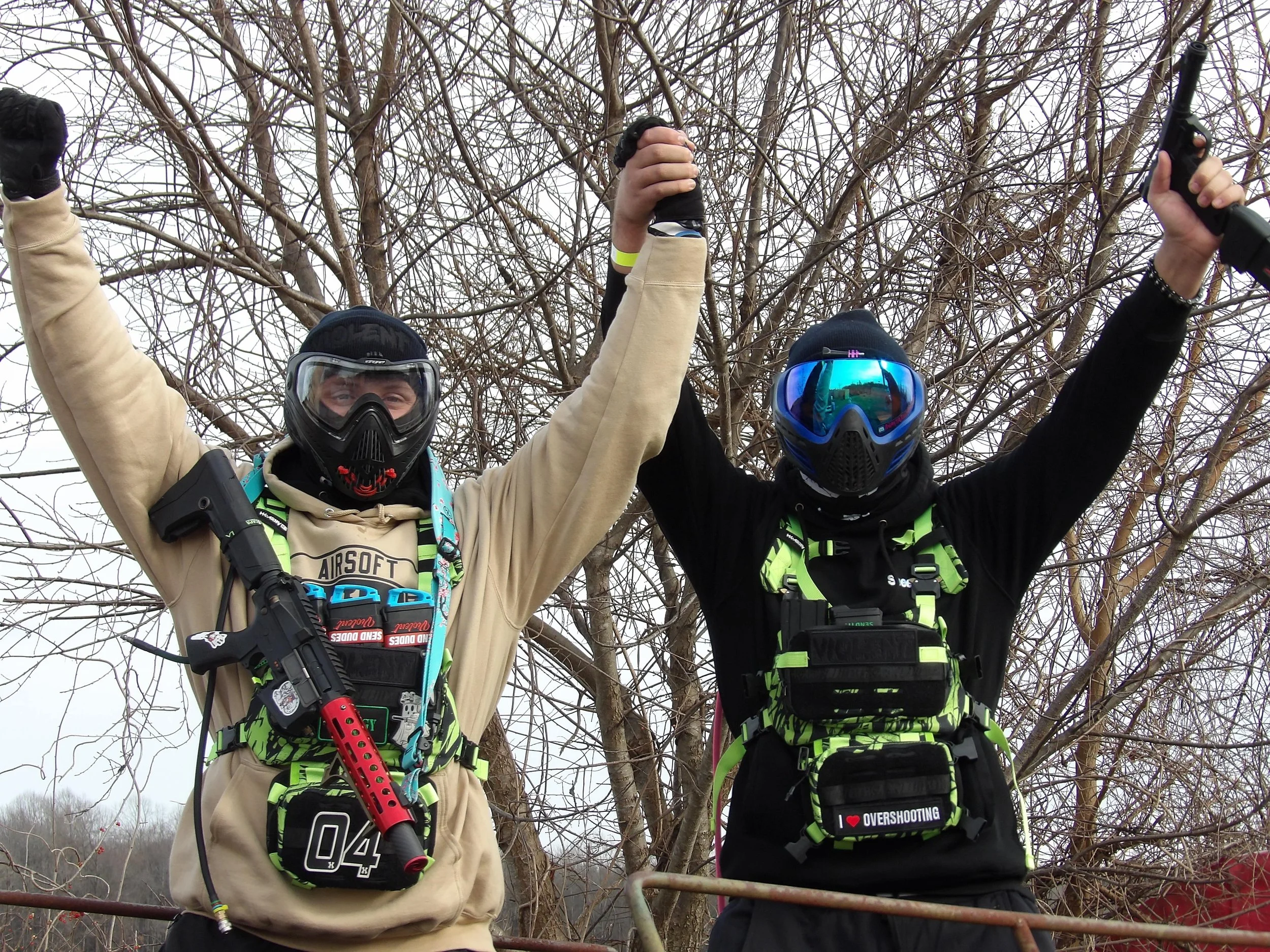 Two individuals wearing protective gear and goggles, raising their arms in celebration outdoors in front of leafless trees, one holding a paintball gun.