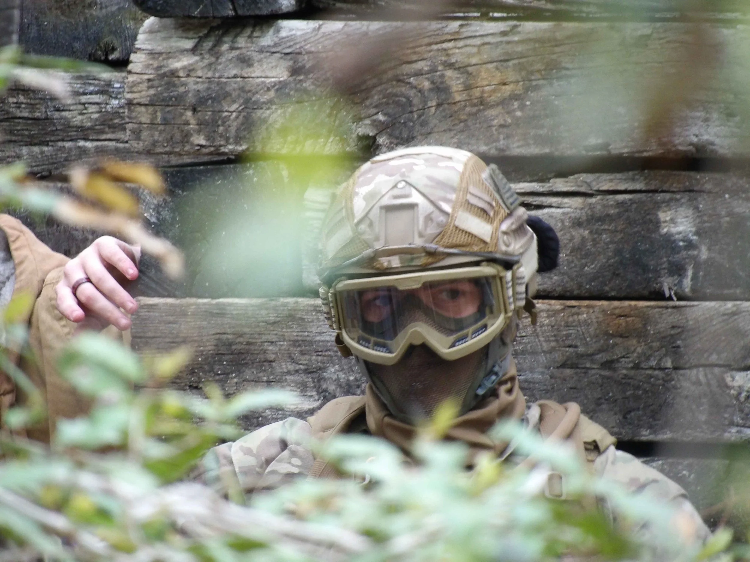 A person wearing camouflage military clothing and tan tactical gear, including protective goggles, is crouching behind bushes near a wooden wall or structure outdoors.