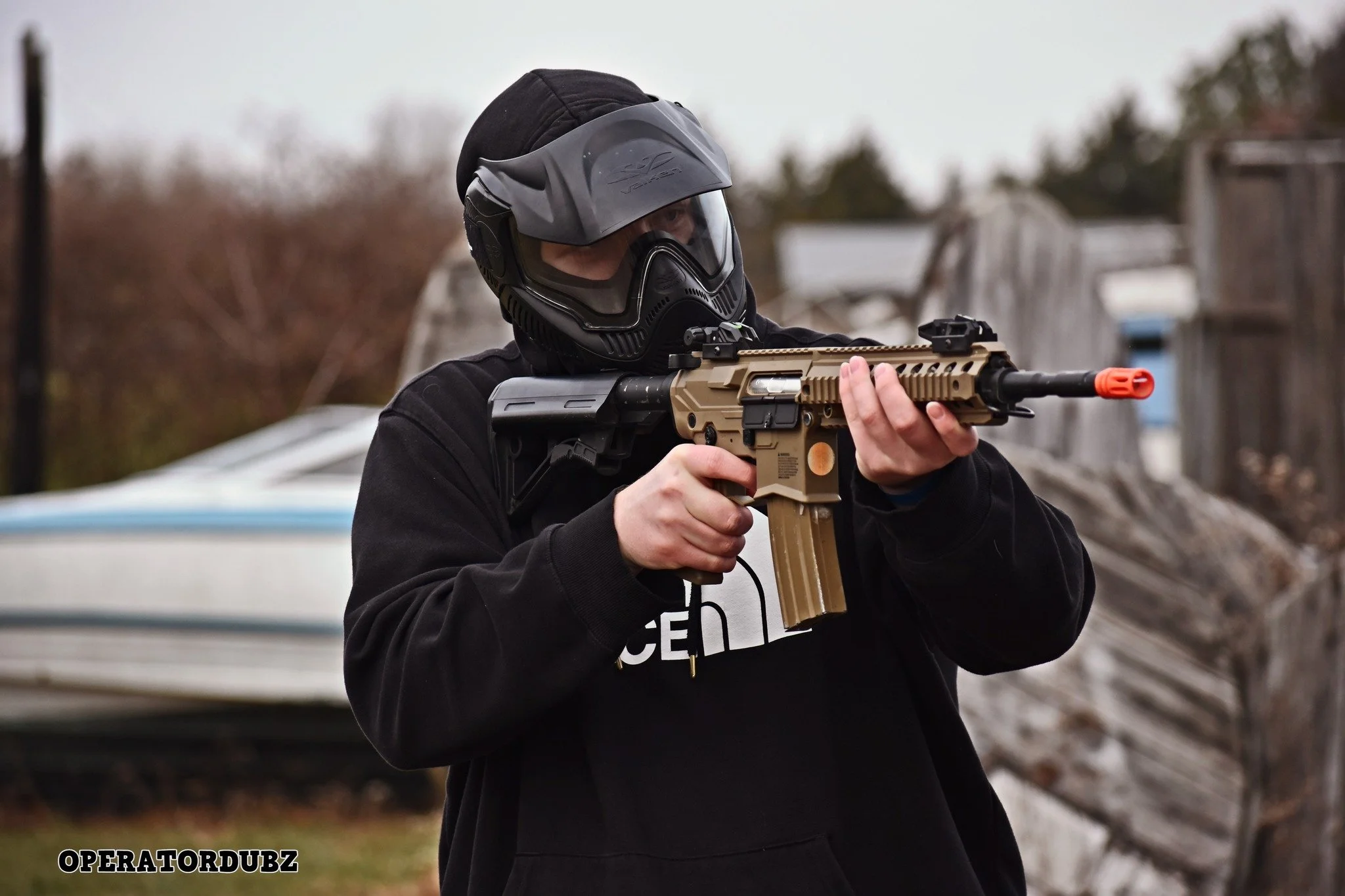 Person wearing a helmet with a visor, aiming a tactical tan and black airsoft or toy rifle outdoors.