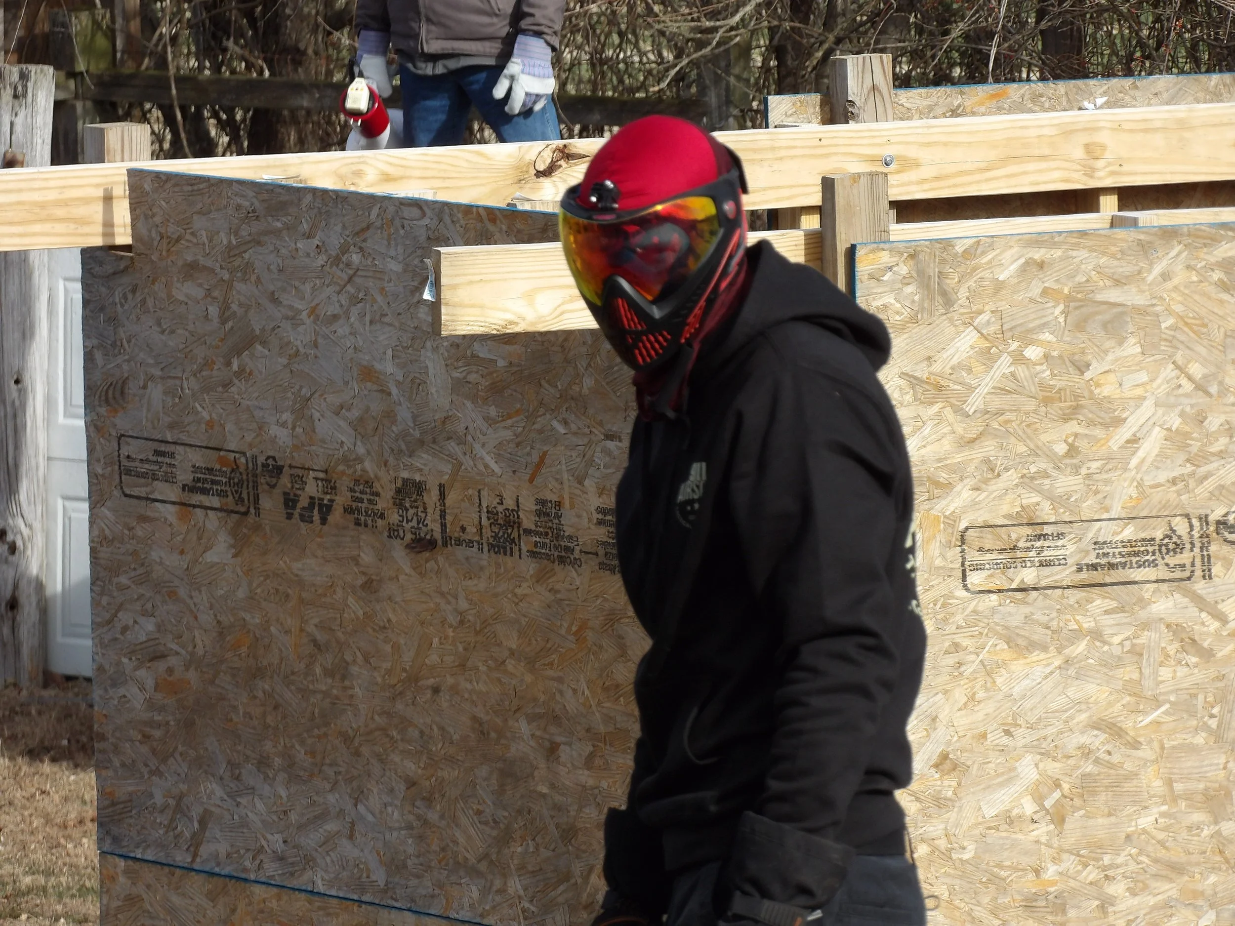 Person wearing black jacket and red face mask with goggles, standing in front of wooden construction wall. Another person in the background wearing gloves and jacket.