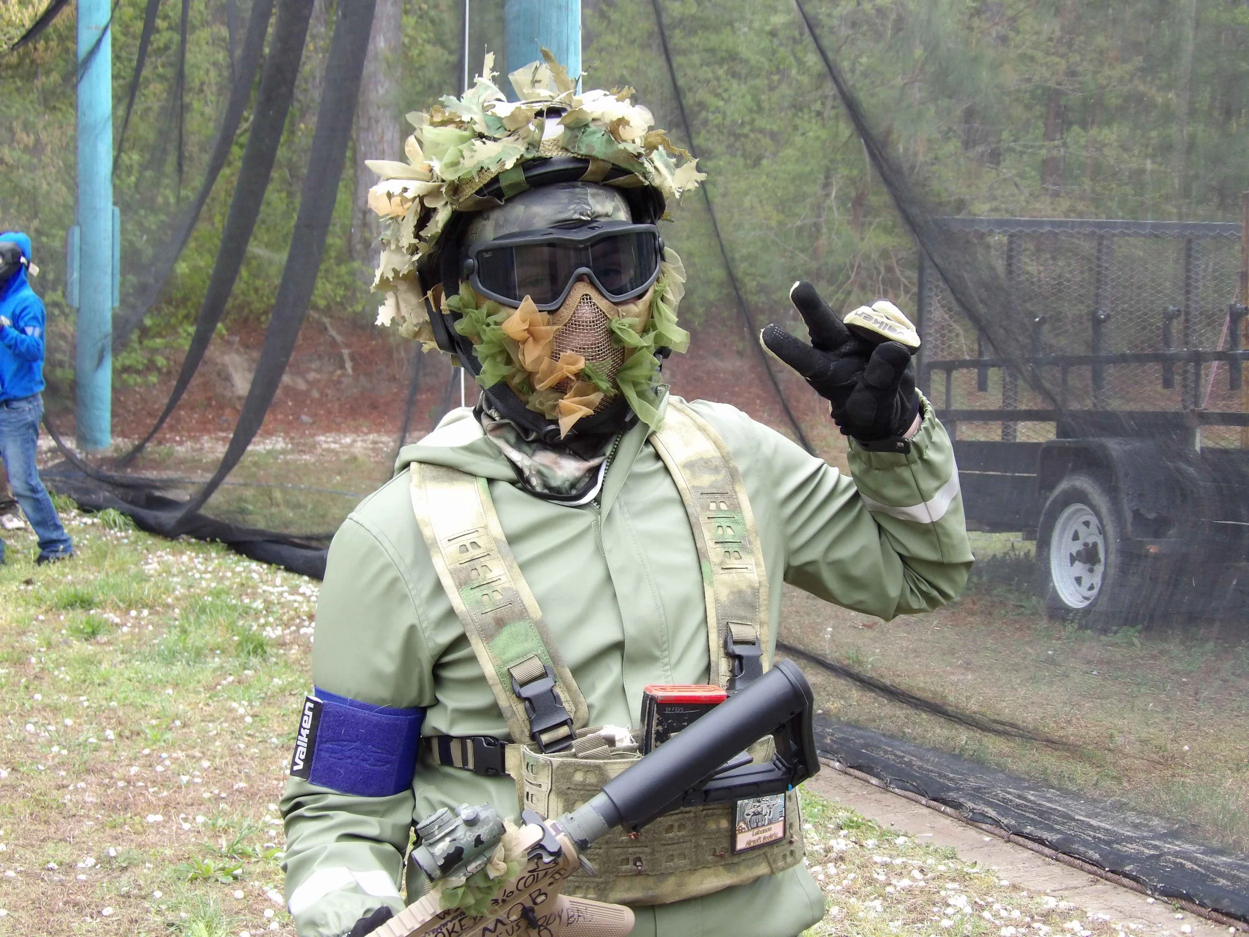 A person dressed in camouflage military gear with a helmet covered in leaves, goggles, and a face mask, giving a peace sign at an outdoor wooded area with a netted fence in the background.