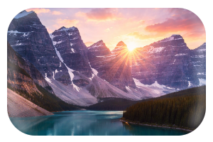 Sunset over a mountain range reflected in a serene lake with a forested shoreline in the foreground.