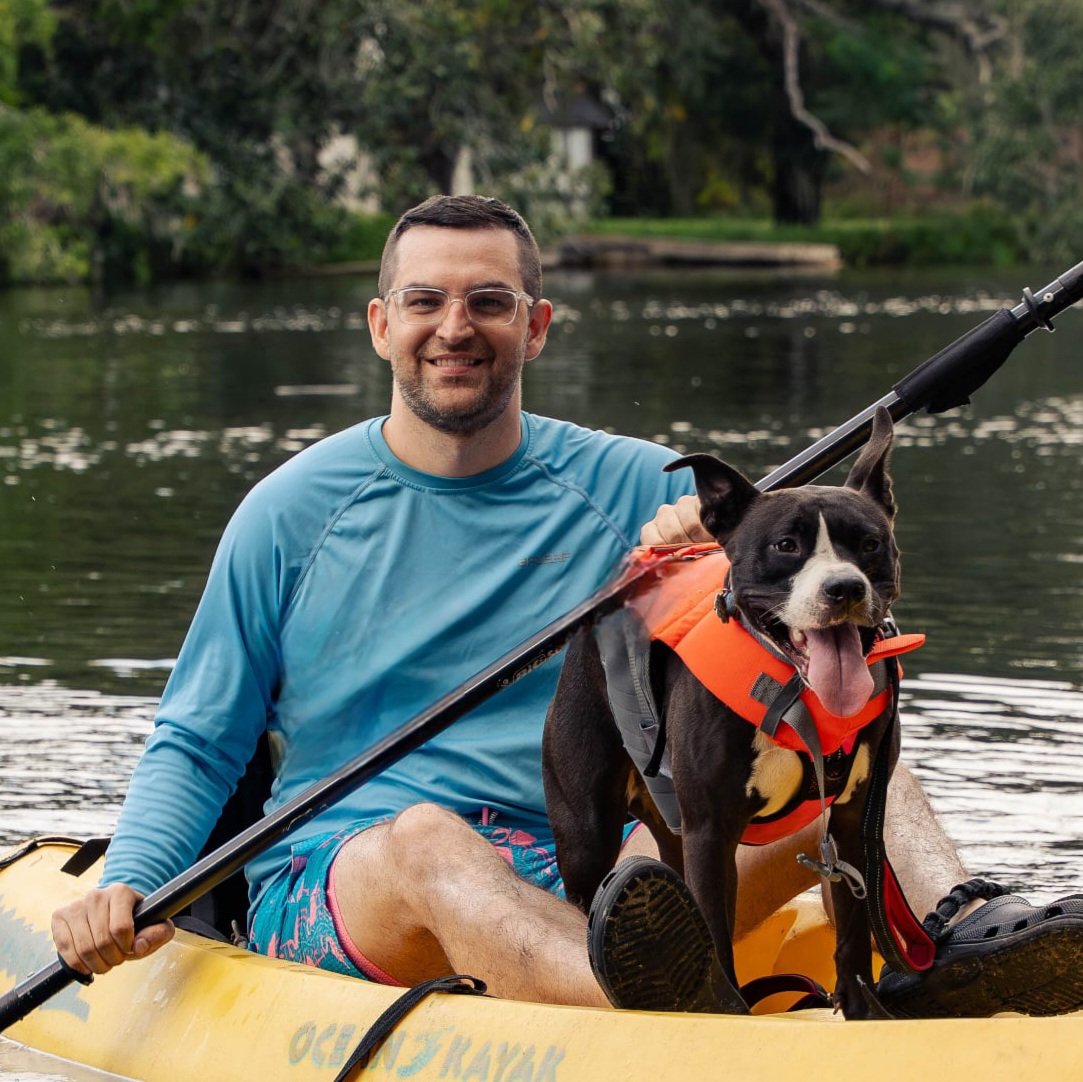 A man and a dog paddle a kayak on a river surrounded by trees, with the man smiling and the dog wearing an orange life jacket and sticking out its tongue.