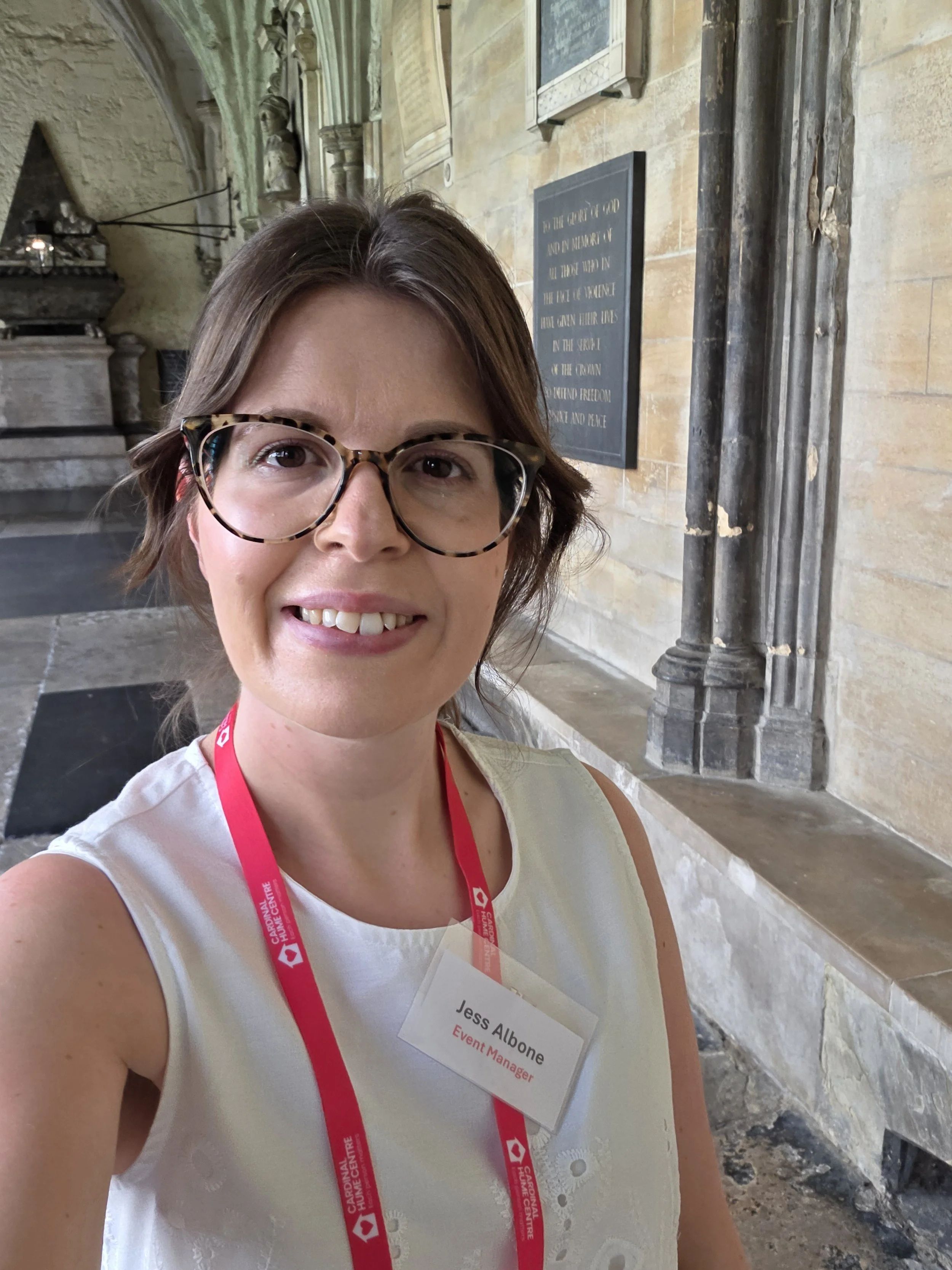 Close-up of a woman with short brown hair, wearing glasses with a tortoiseshell pattern, smiling at the camera. She has a name badge that reads 'Jess Albone' and 'Event Manager,' and wears a lanyard. The background shows a historic stone building with plaques and ornate architectural details.
