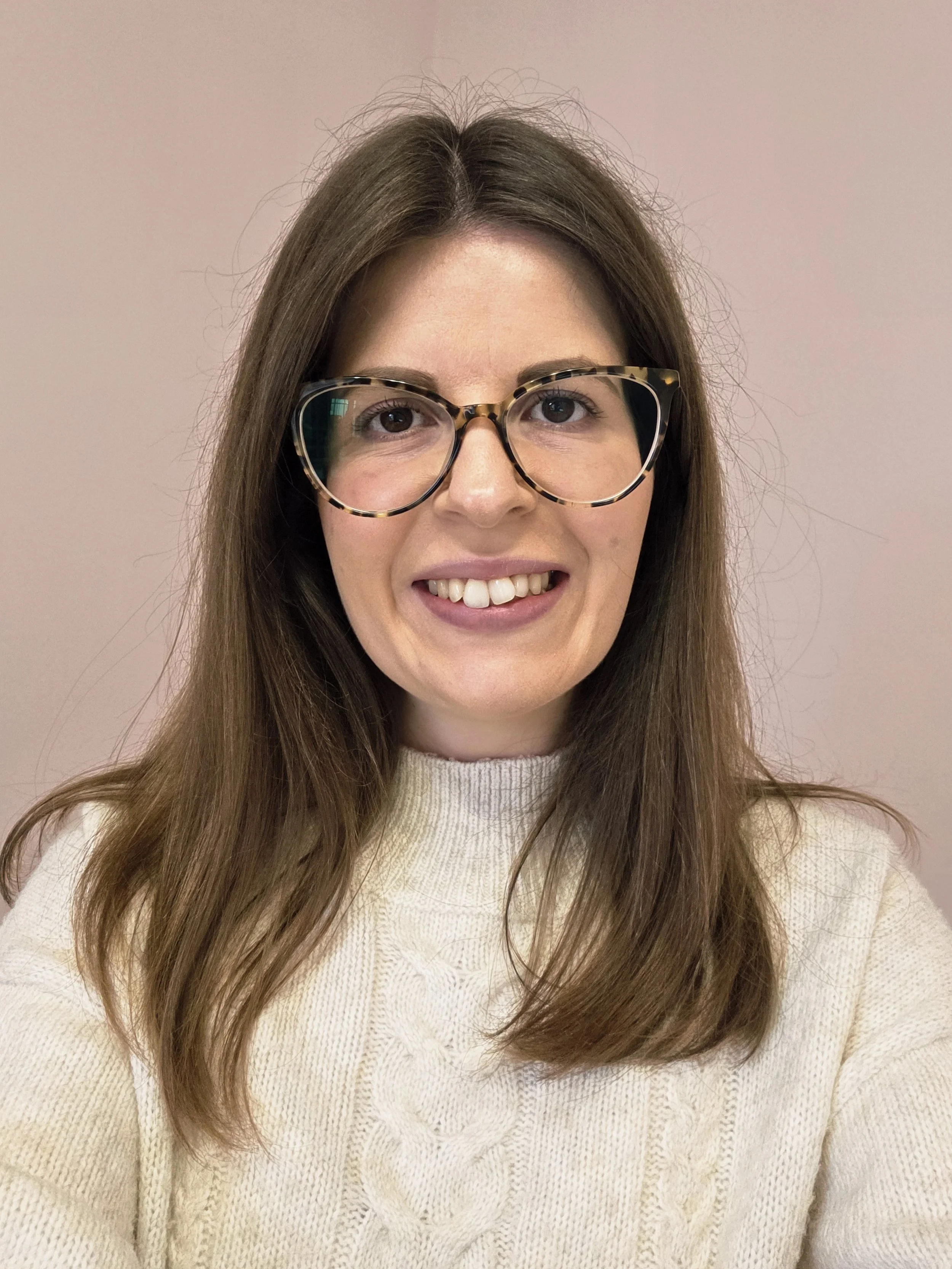 A woman with long brown hair wearing large tortoise shell glasses and a cream sweater, smiling in front of a plain pink wall.