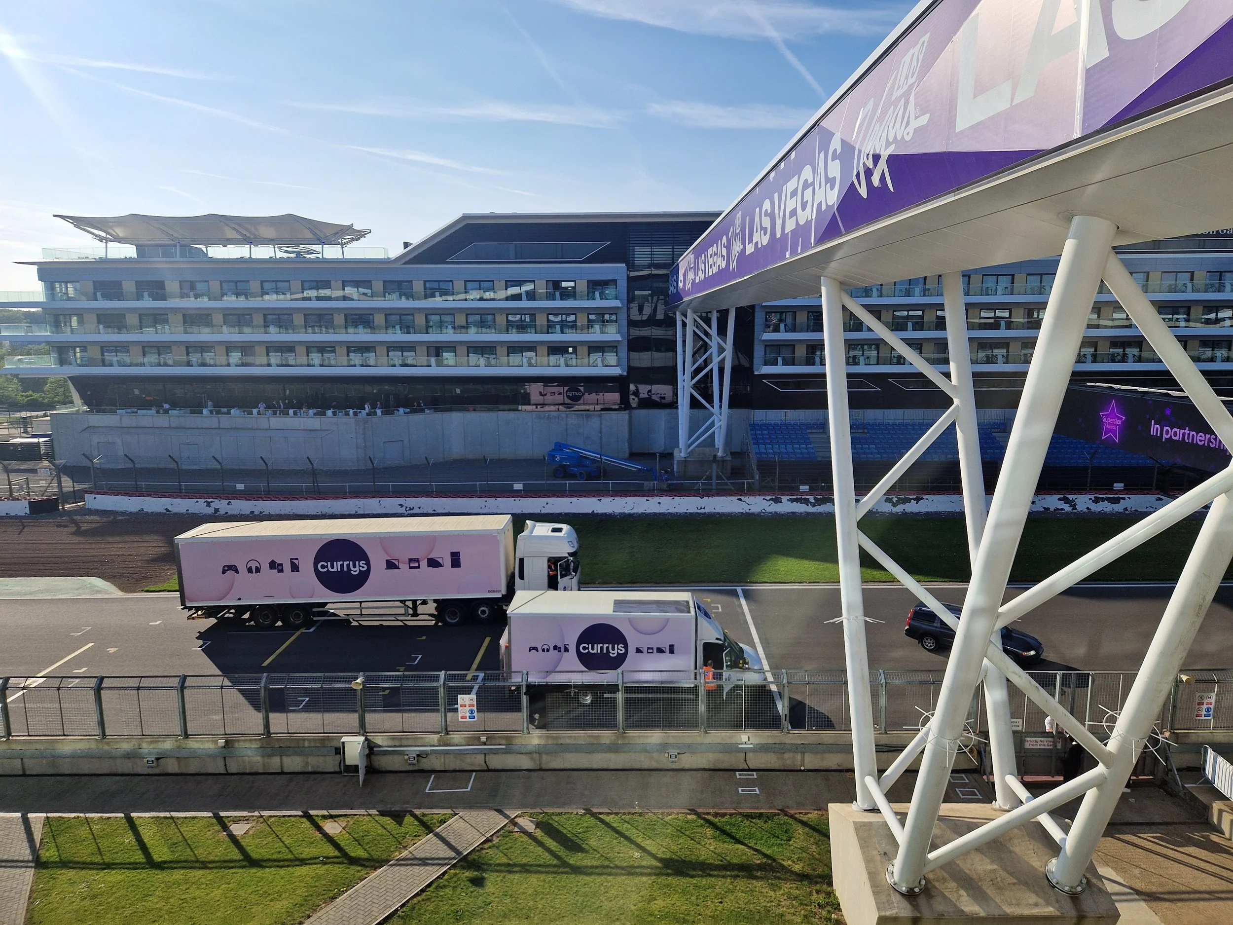 View of a modern outdoor event venue with a multi-story building, purple banners displaying 'Las Vegas' and 'In partnership with', delivery trucks labeled 'Currys', and a sporting track in the foreground.
