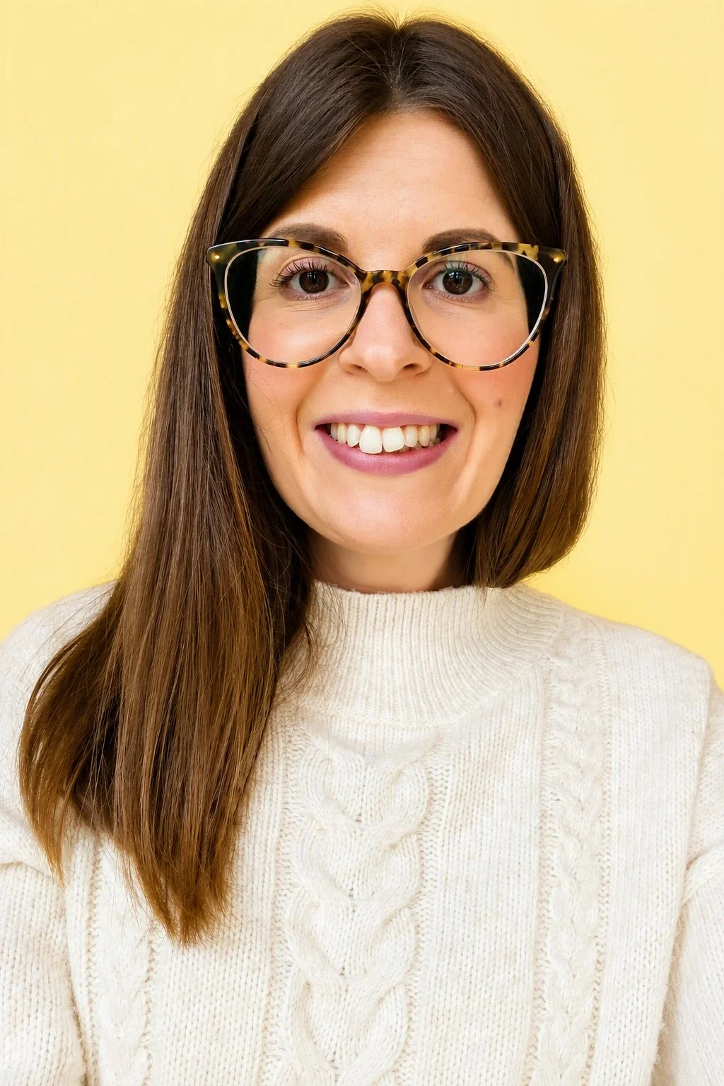 A woman with long brown hair, wearing large tortoiseshell glasses and a cream-colored cable-knit sweater, smiling against a yellow background.