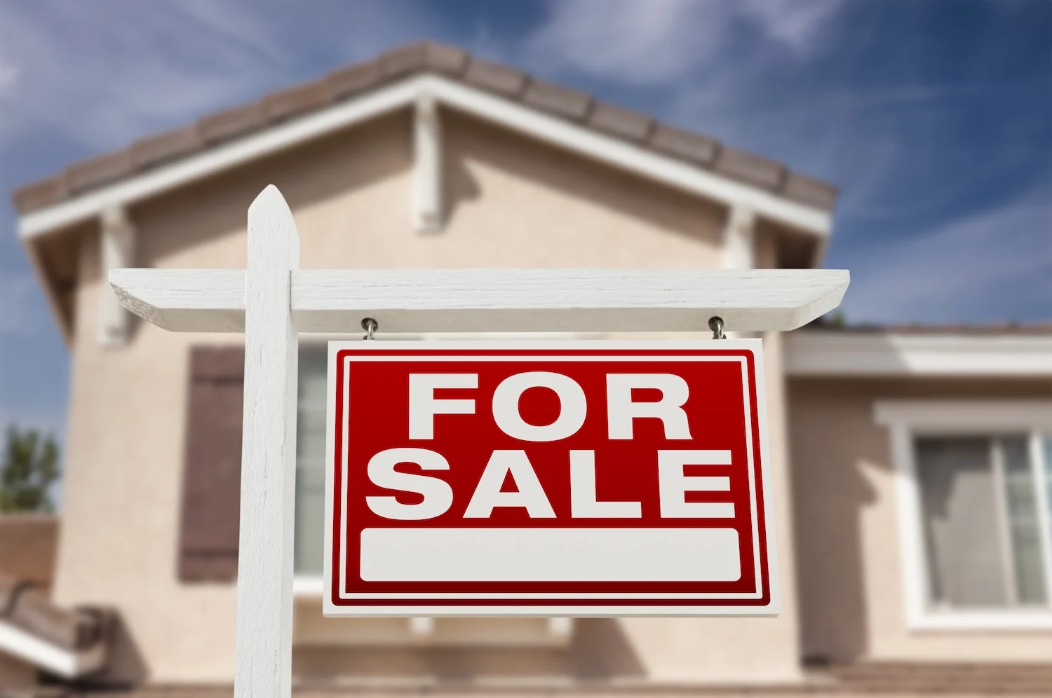 A red and white 'For Sale' sign hanging on a white wooden post in front of a beige house with a brown tiled roof.