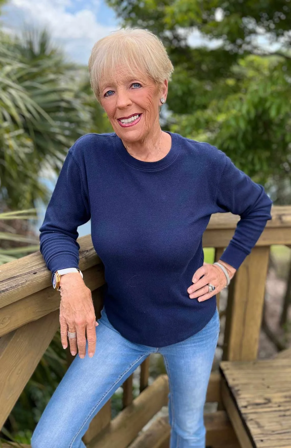 A woman with short blonde hair, blue eyes, wearing a dark blue long sleeve top and light blue jeans, smiling, standing outdoors on a wooden structure with green trees in the background.