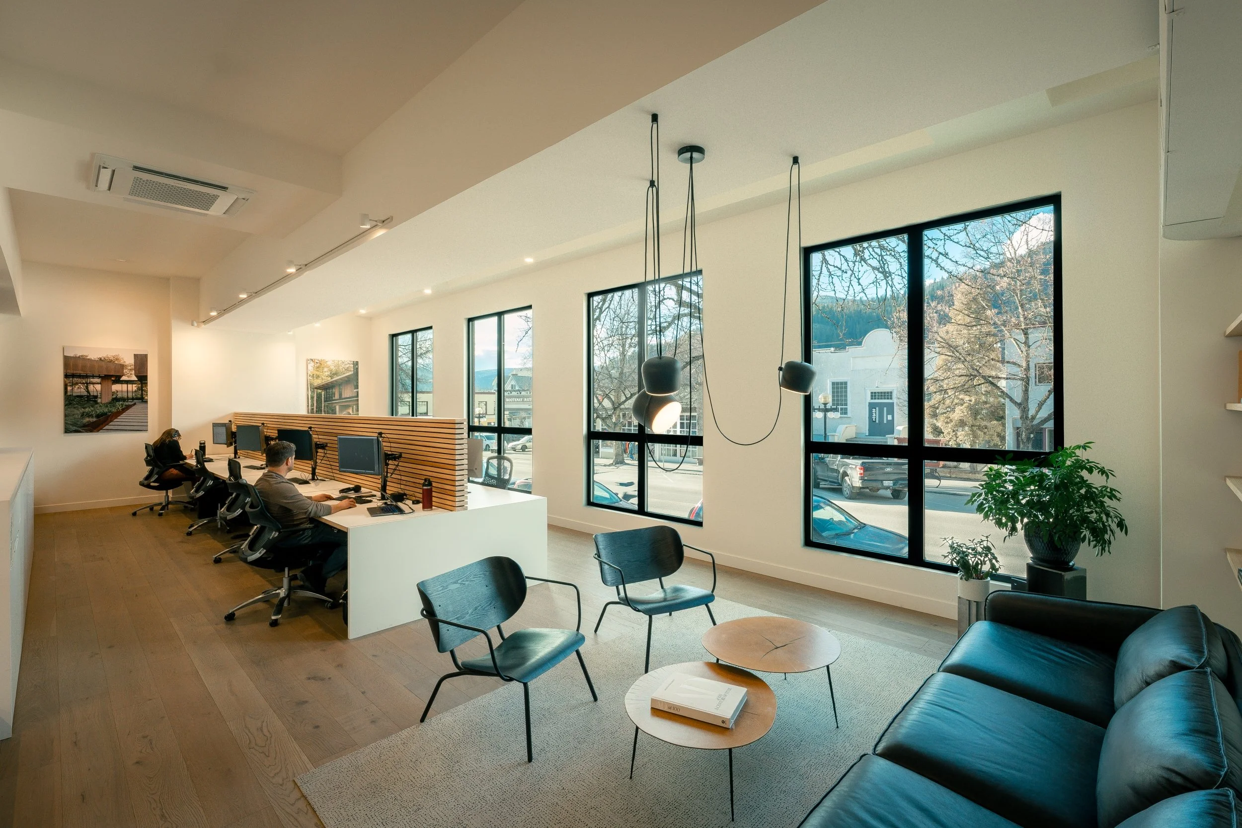 Interior of a modern office with large windows, two people working at desks, sitting on ergonomic chairs, a waiting area with black chairs and a coffee table, and a leather sofa near a potted plant.