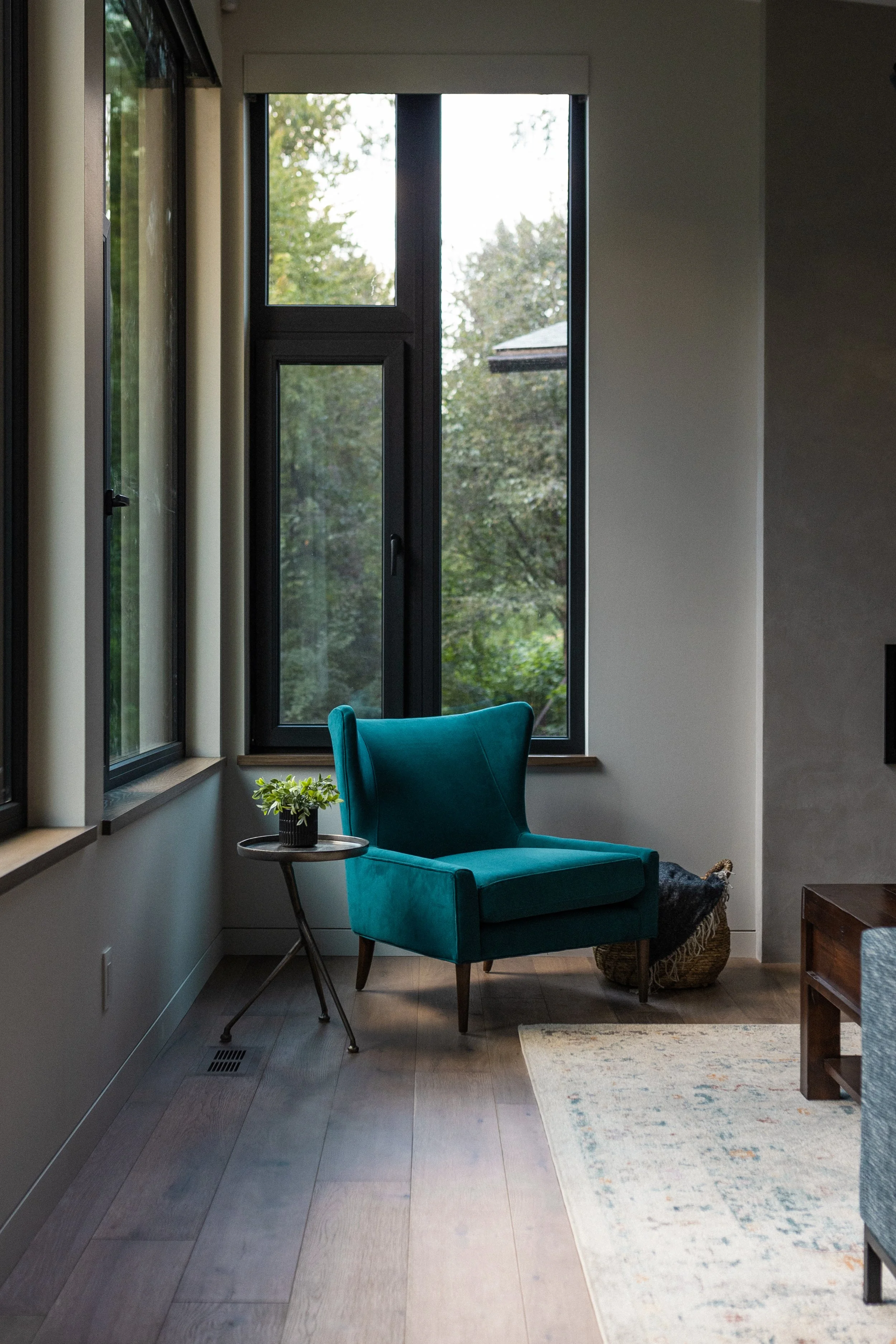 Modern living room with a teal armchair, side table, potted plant, large black-framed windows, and trees visible outside