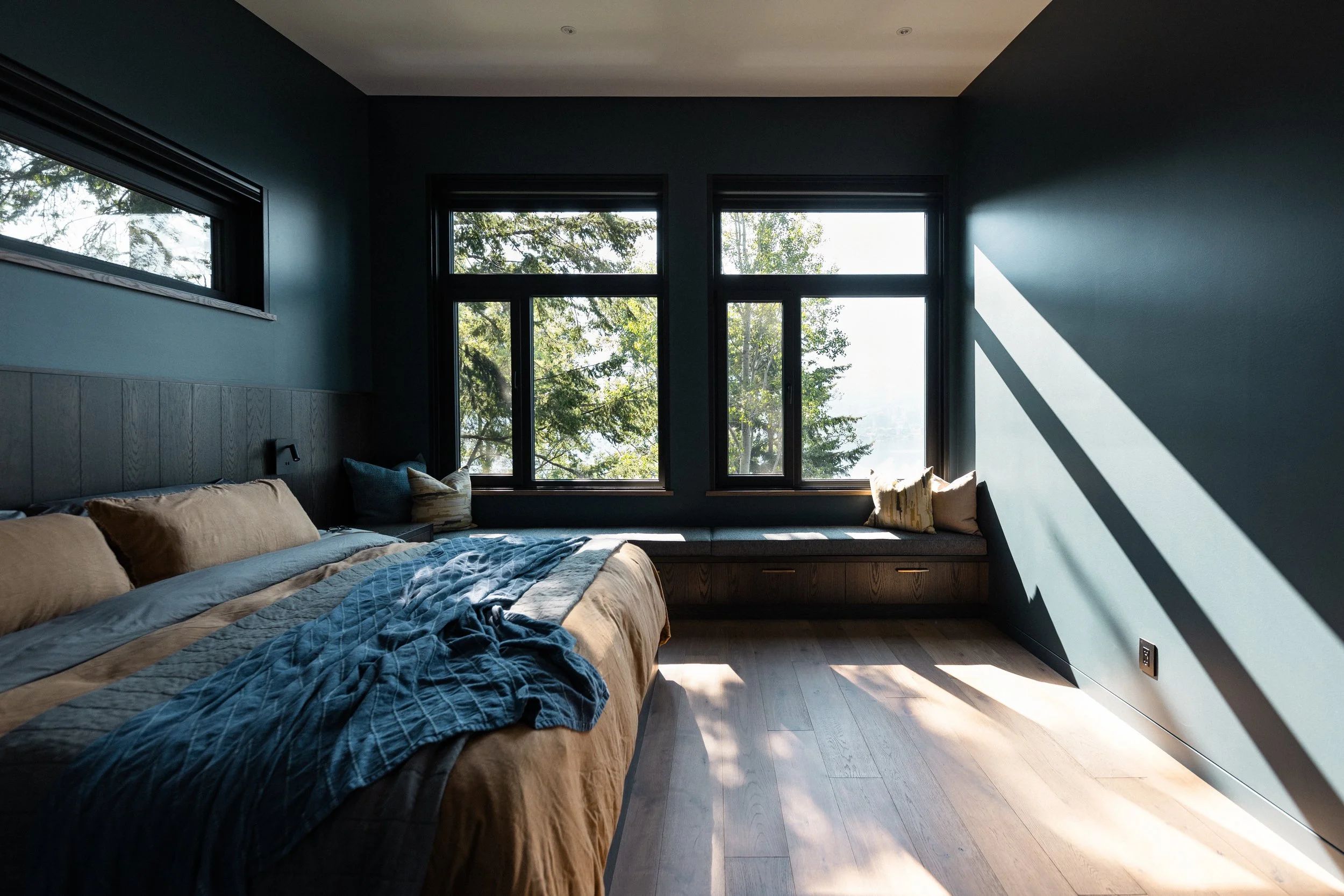 Modern dark bedroom with a neatly made bed, built-in window seat, large black-framed windows, and sunlight casting shadows across the wood floor.