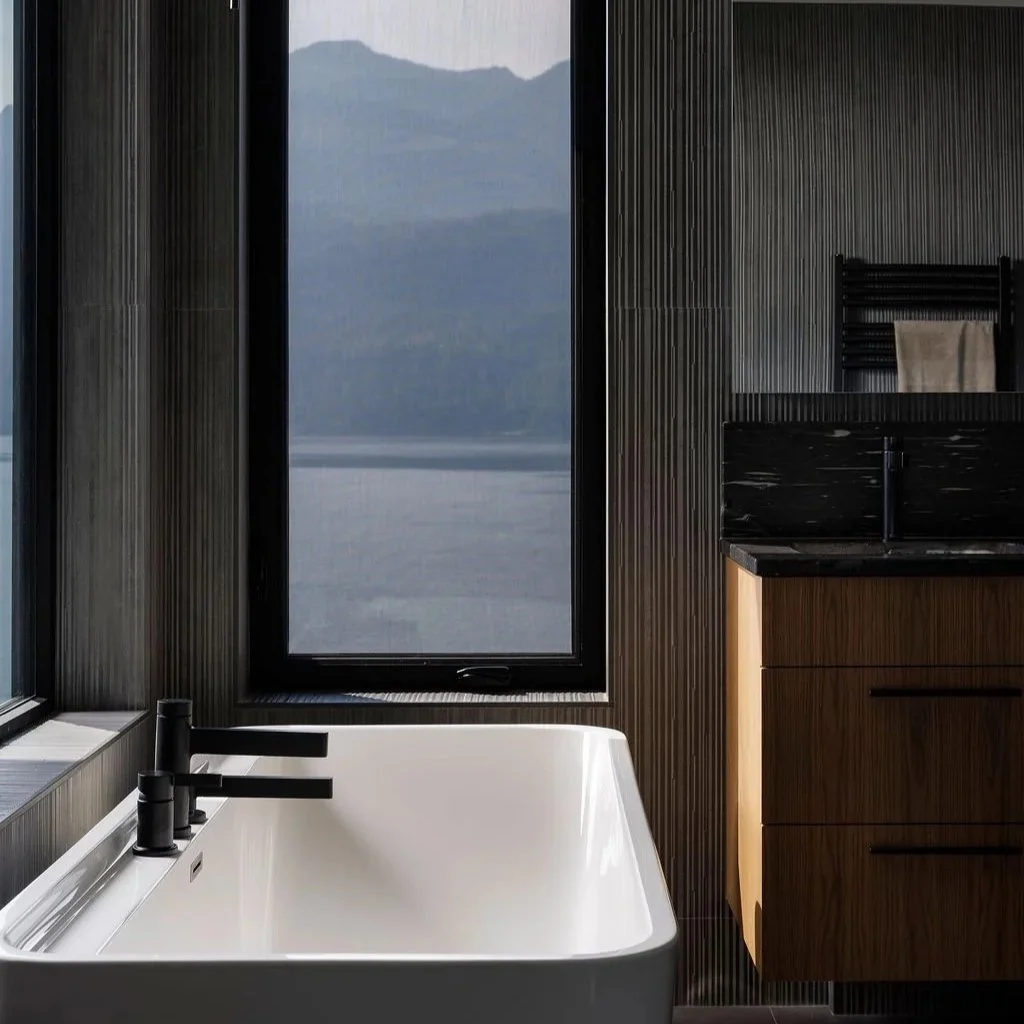 Modern bathroom with a white bathtub, black faucet, wooden cabinet, large window with mountain view, black textured wall, and a black and white pendant light.