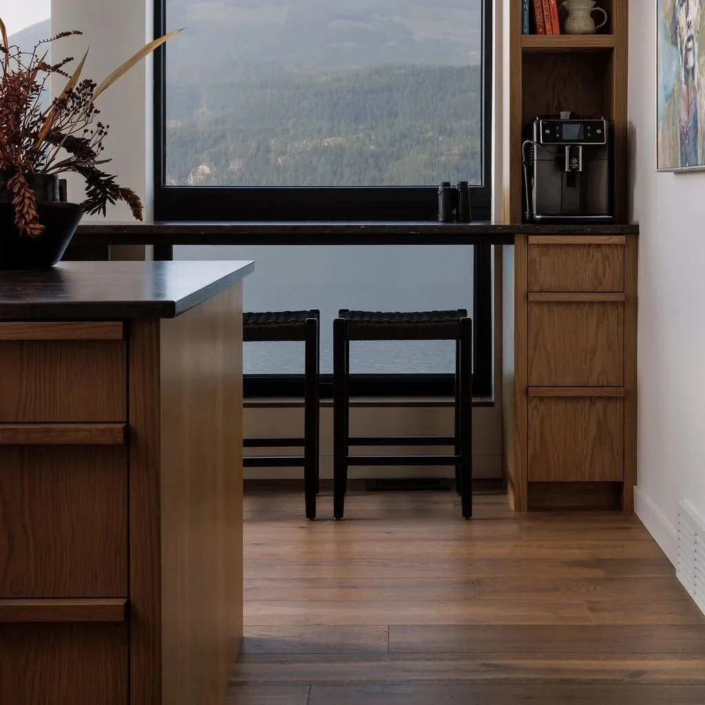 A kitchen with a large window showing a mountainous landscape outside. The kitchen features wooden cabinetry, a black countertop with a coffee machine, and open shelves with cups, bowls, and books. Two black chairs are placed in front of the window.