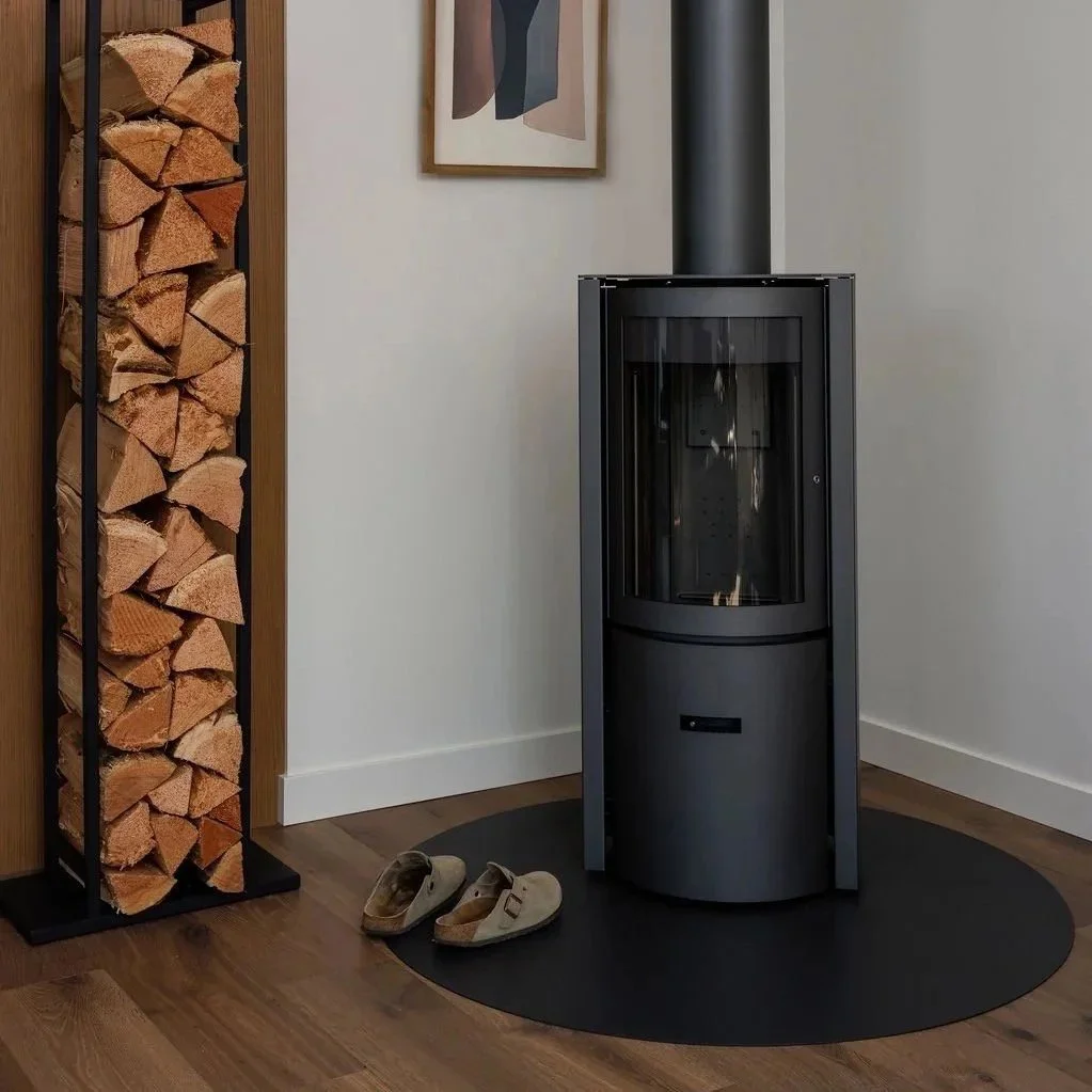 A wood-burning stove with a black chimney pipe in a corner of a room, next to a wood log storage rack and a framed abstract art piece on the wall, with a pair of shoes on a circular black rug on the wooden floor.