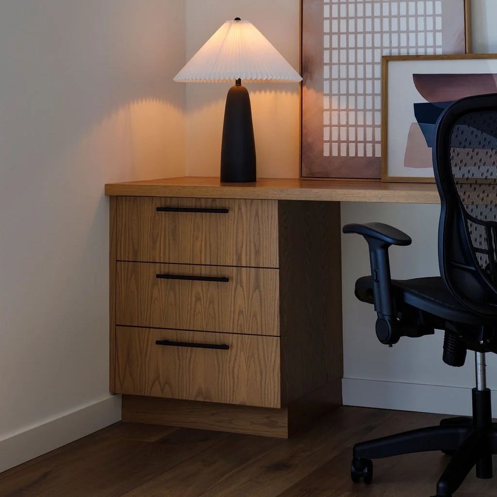Corner of a modern home office with a wooden desk, black office chair, a black bedside lamp with a white lampshade, and framed wall art, including abstract prints and patterned art, against a white wall.