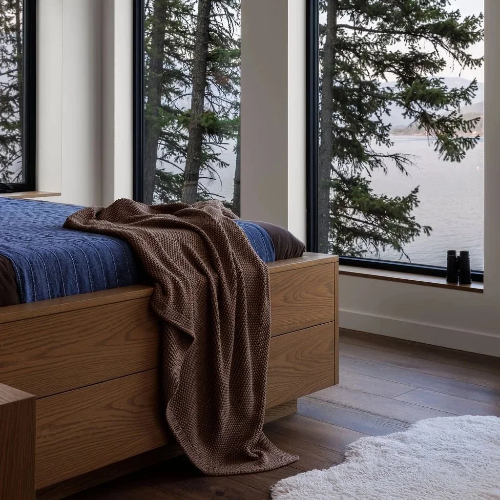 A bedroom with large windows overlooking a lake and tall trees outside. The bed has a blue quilt and a brown blanket, with a white rug on the wooden floor.