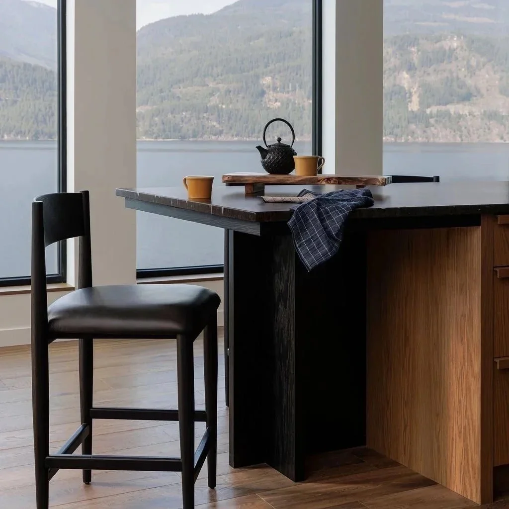 Interior of a modern kitchen with large windows overlooking mountains and a lake, a black teapot, yellow cups, a wooden cutting board, and a dark counter with a black chair.