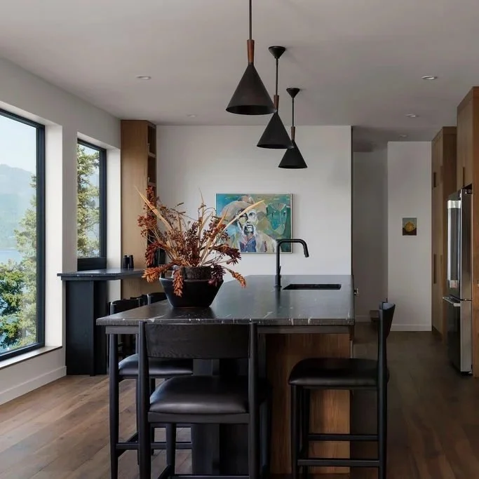 Modern kitchen with large window showing trees outside, black pendant lights over a black island with a vase of dried flowers, built-in wooden cabinets, stainless steel appliances, and a wood storage unit.