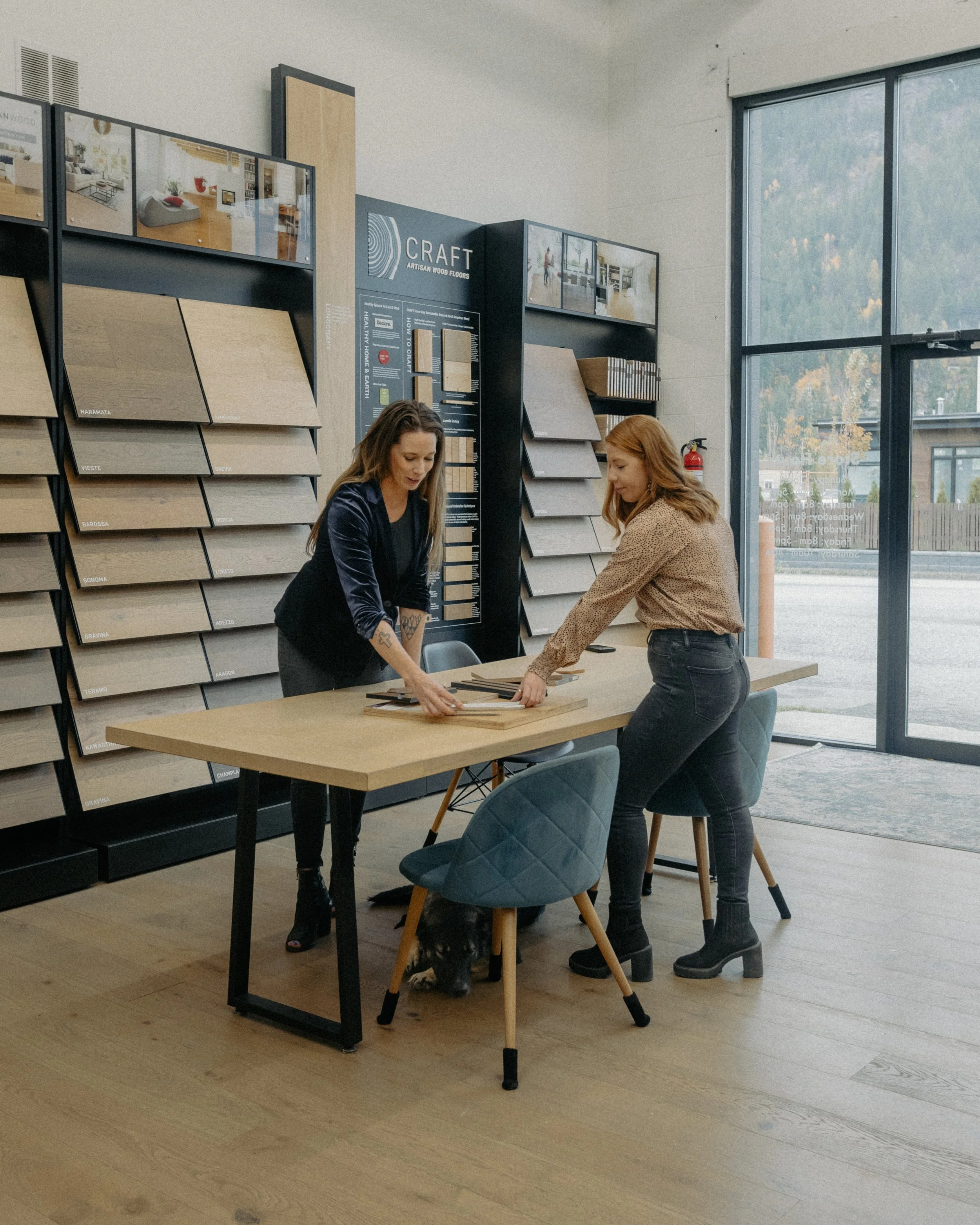 Two women examine wood flooring samples in a flooring showroom with wall displays and a dog beneath the table.