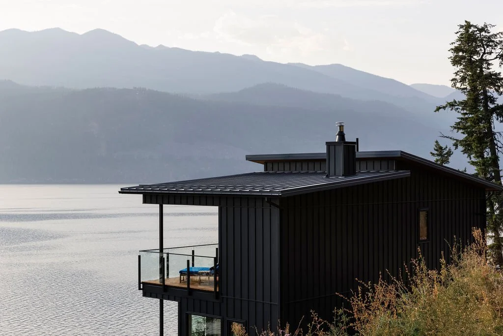 A black modern house with a balcony overlooking a lake, with mountains in the background and trees on the right side.