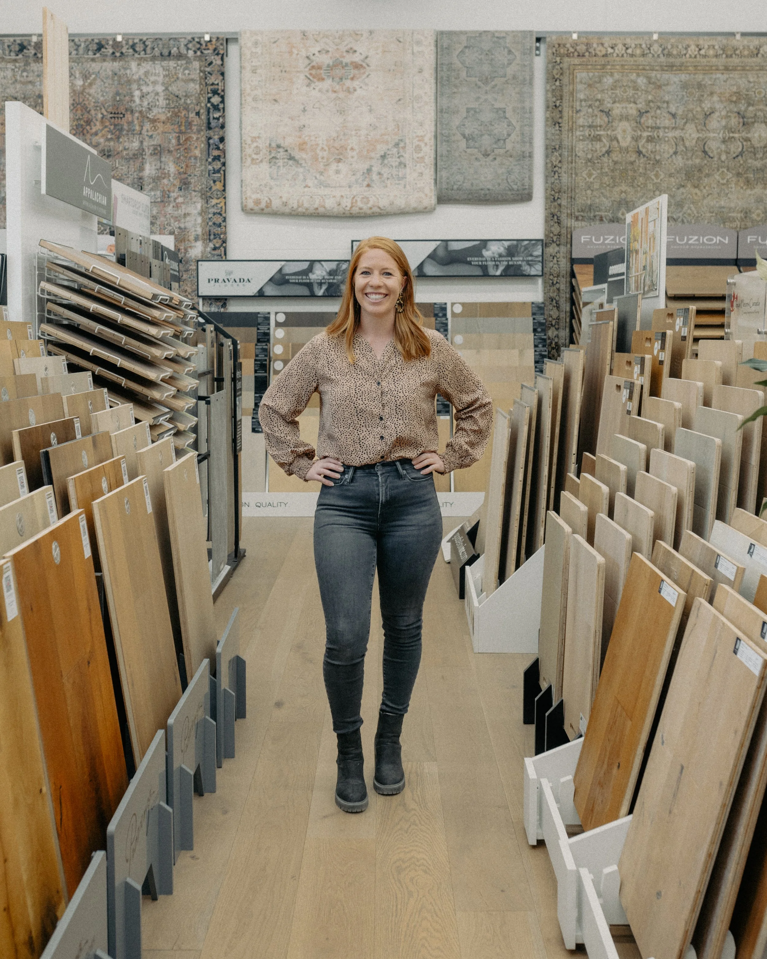 A woman with red hair and a beige polka-dot shirt standing with hands on hips in a flooring store aisle, smiling. Surrounding her are display racks of wood and tile flooring samples, with rugs hanging on the wall in the background.