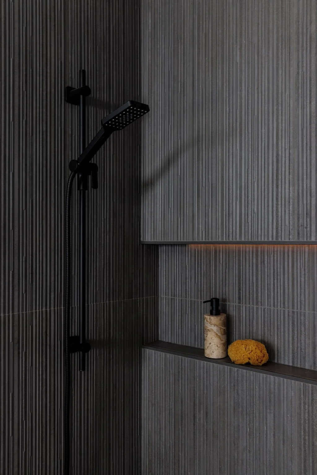 Dark tiled shower with black showerhead, shelf holding a soap dispenser and a yellow sponge.