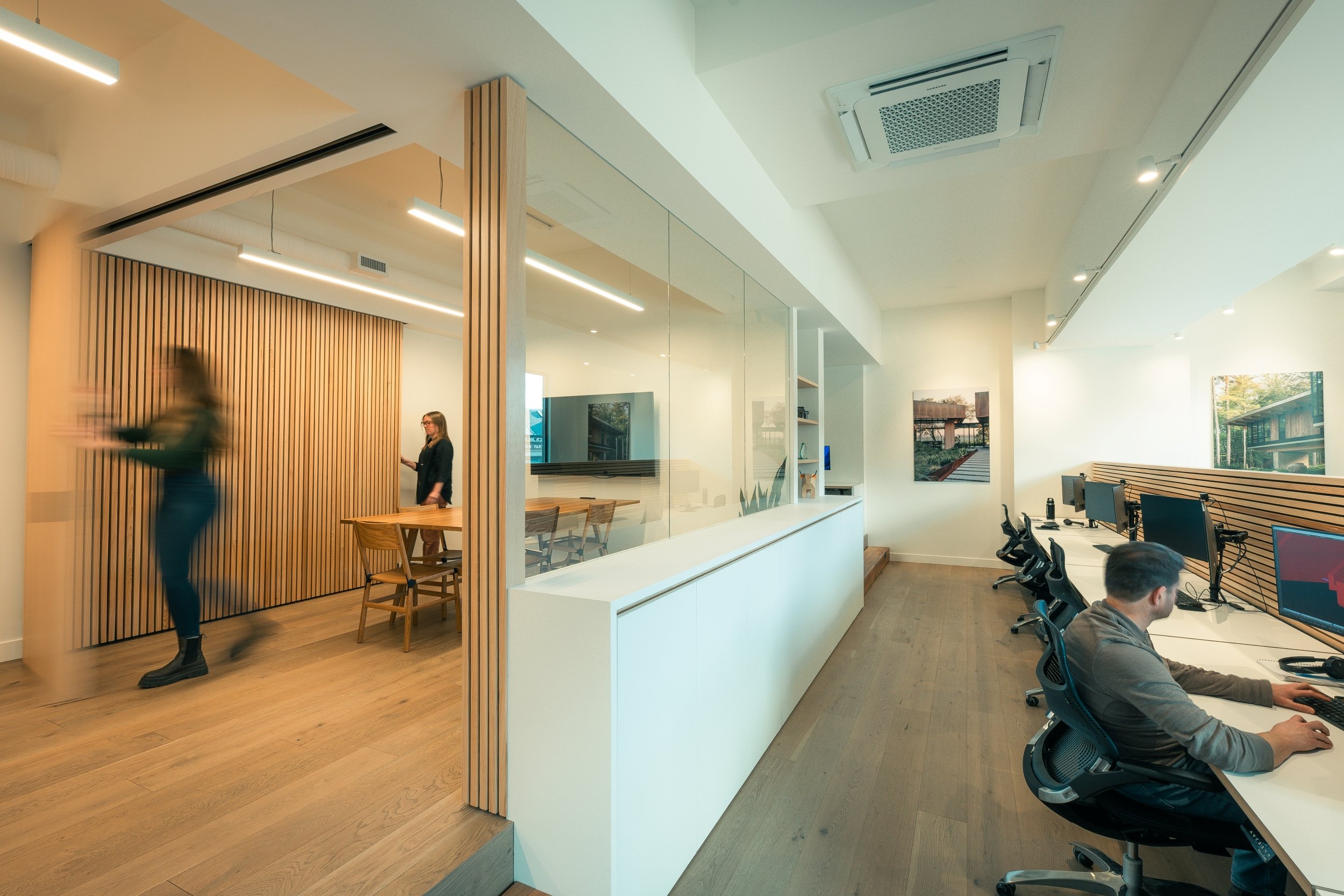 Modern office space with people working at desks, wooden room divider, wall with artwork, large windows allowing natural light, mounted TV, and ceiling air vents.