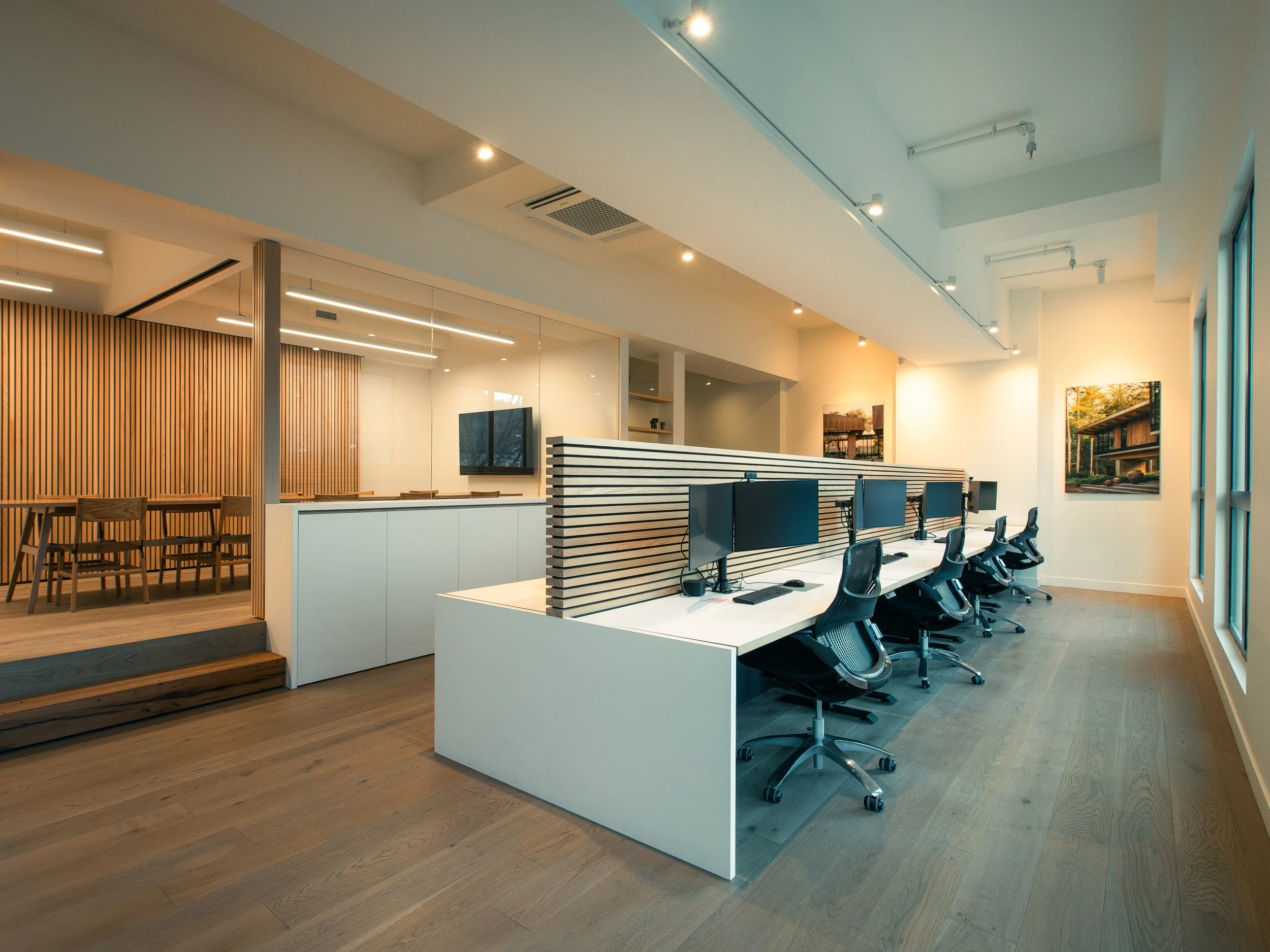 Modern office space with a row of computer desks and black office chairs, separated by a wooden slat divider, with artwork on the wall and large windows letting in natural light.