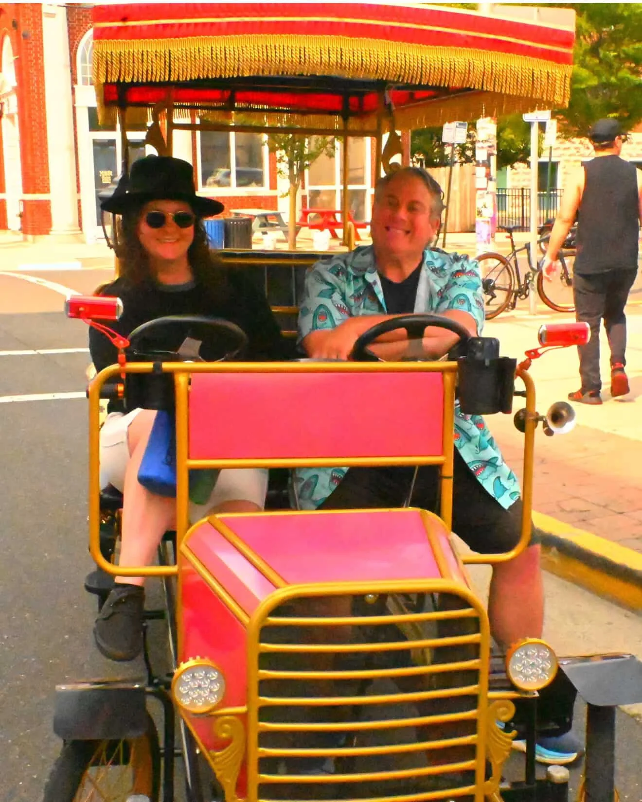 A man and woman riding a colorful four-wheeled pedal vehicle with a canopy, smiling and enjoying the ride in an urban setting with buildings, bicycles, and pedestrians in the background.