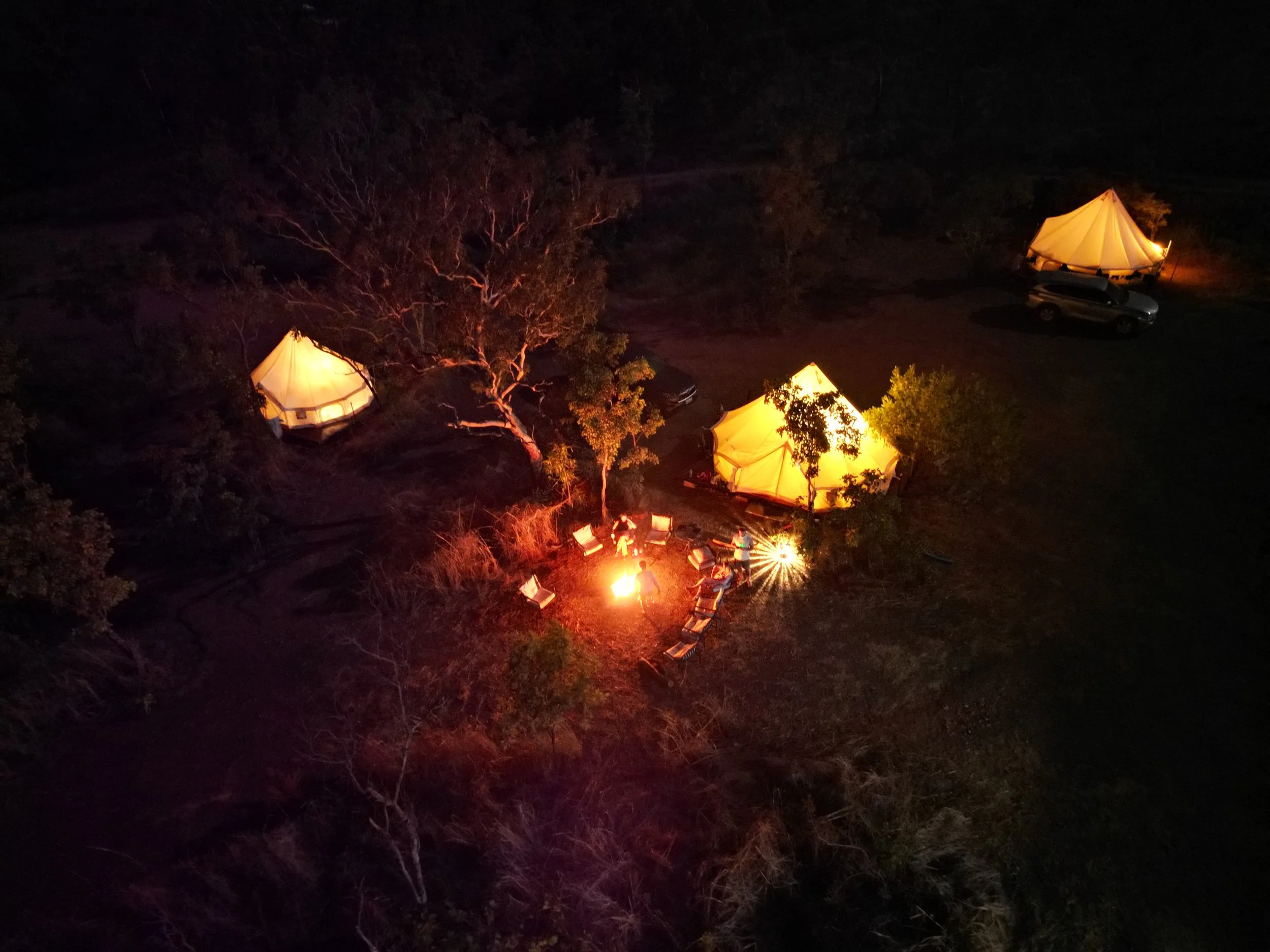 Nighttime outdoor scene with illuminated tents and a campfire, surrounded by trees.